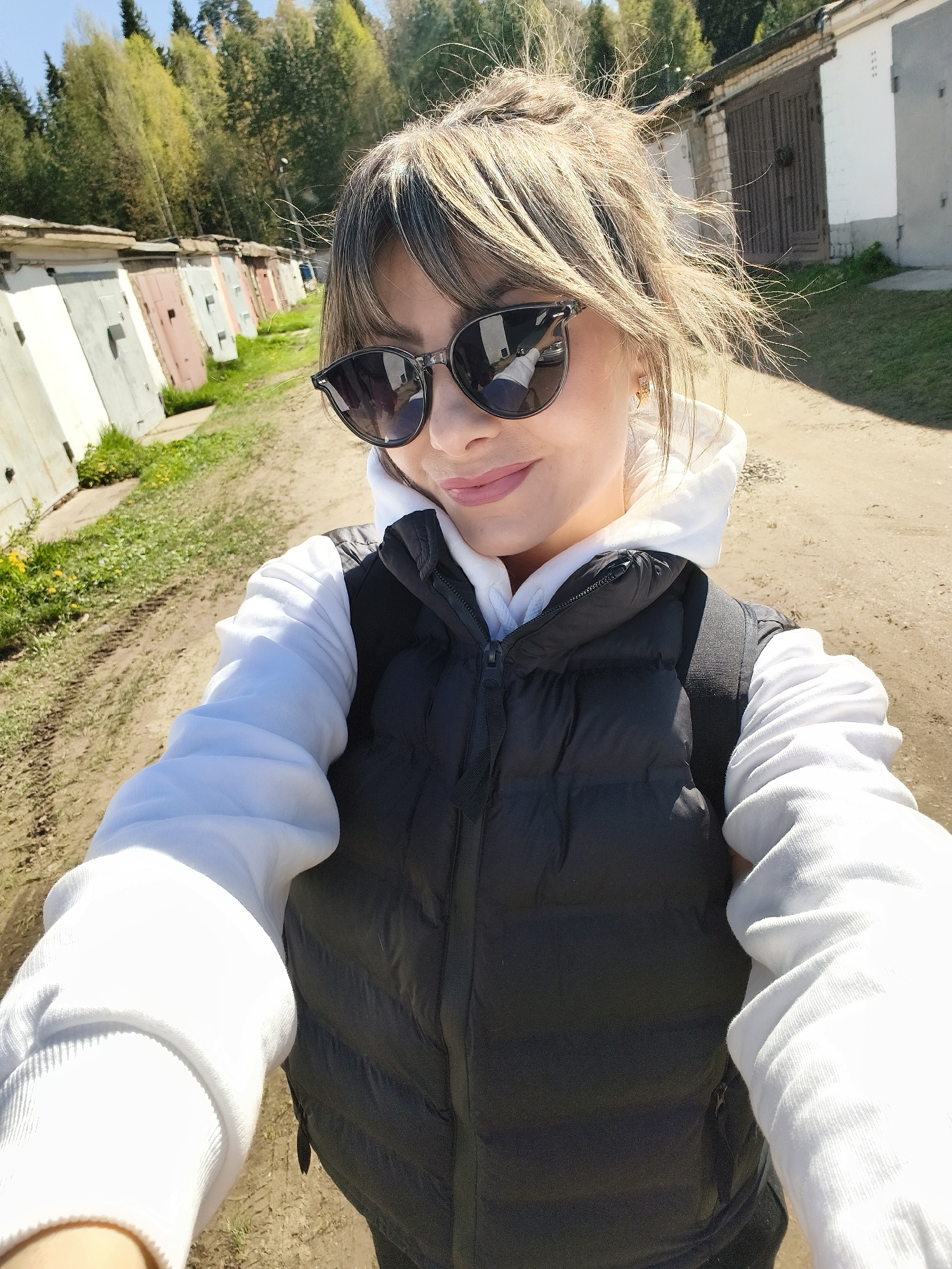 The image shows a woman taking a selfie. She is wearing sunglasses, a white hoodie, and a black vest. She is standing on a dirt road in what appears to be an industrial area, with a row of garages visible on the left and green foliage on the right. Based on the location information, the image was likely taken in Chaikovsky, Russia. The environment suggests a bright, sunny day.