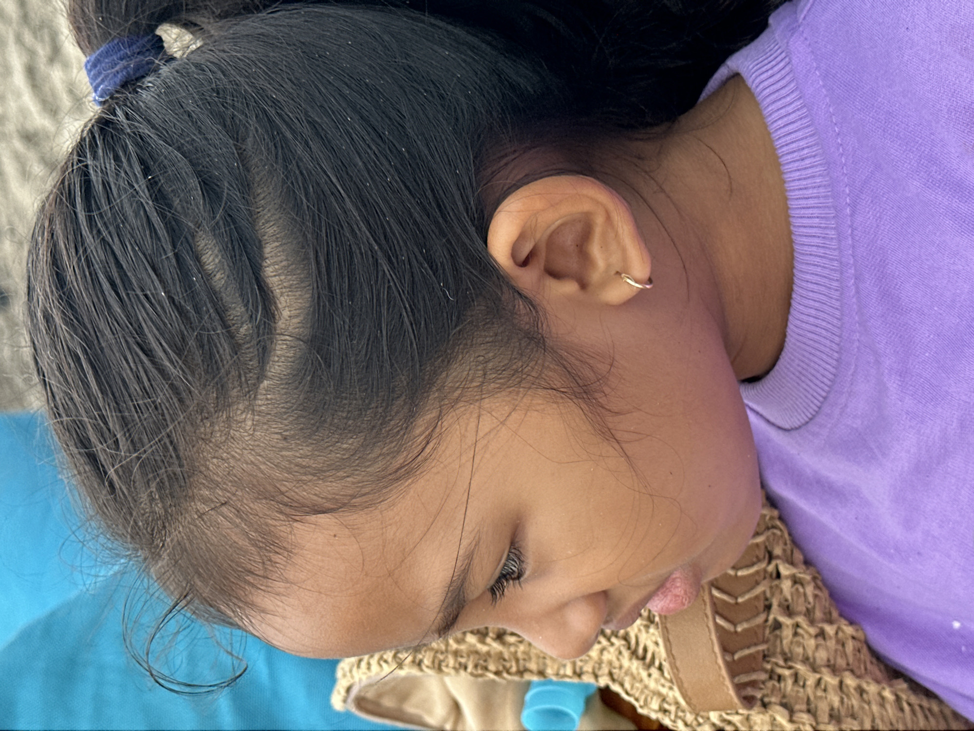 A medium shot, profile view of a young Southeast Asian girl is presented, focusing on her head and upper body. She has dark hair pulled back into a ponytail with a blue hair tie and is wearing a purple crewneck shirt. A thin gold hoop earring is visible in her left ear. To the left of the girl, a portion of a straw bag is visible, with a bright blue object protruding from it. The background is blurred, with hints of sand and a blue fabric. The image appears to have been taken outdoors, possibly at a beach in North Kuta, Indonesia.