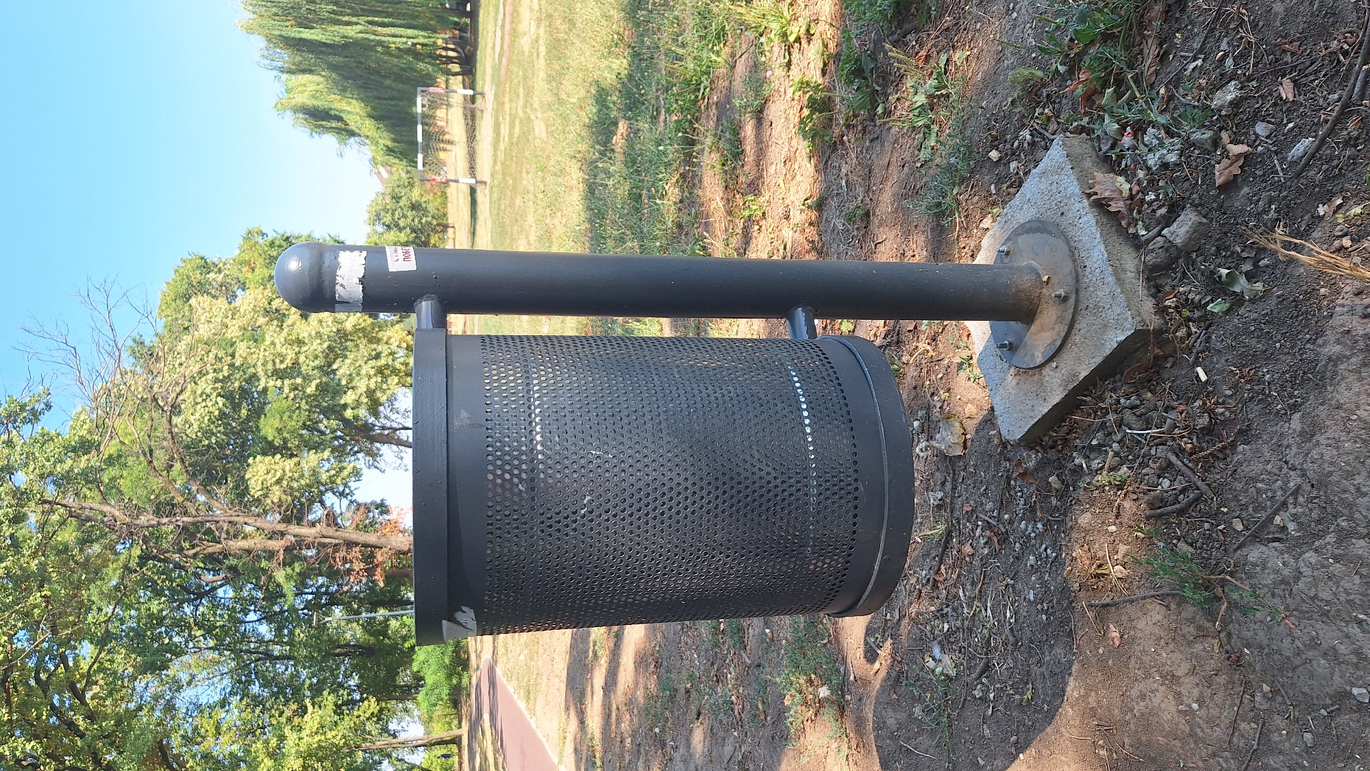 The image depicts a public park scene on a sunny day. In the foreground, a black, perforated cylindrical trash bin is mounted on a dark gray pole, which is anchored to a concrete base. A small, white sticker with Cyrillic text is visible on the pole near the top. The trash bin is positioned on a dirt path with scattered dry leaves and twigs.
Behind the trash bin, a red running track curves to the left. Further back, a grassy area stretches towards a goalpost of a soccer field, with a large weeping willow tree dominating the right side of the background. Several mature deciduous trees with green and light-colored foliage are visible in the mid-ground and background, suggesting a park environment. The sky is clear and blue.
This scene is consistent with a park located in Sombor, Serbia, a city known for its numerous green spaces and recreational areas. The presence of a running track and a soccer field further supports this identification.
