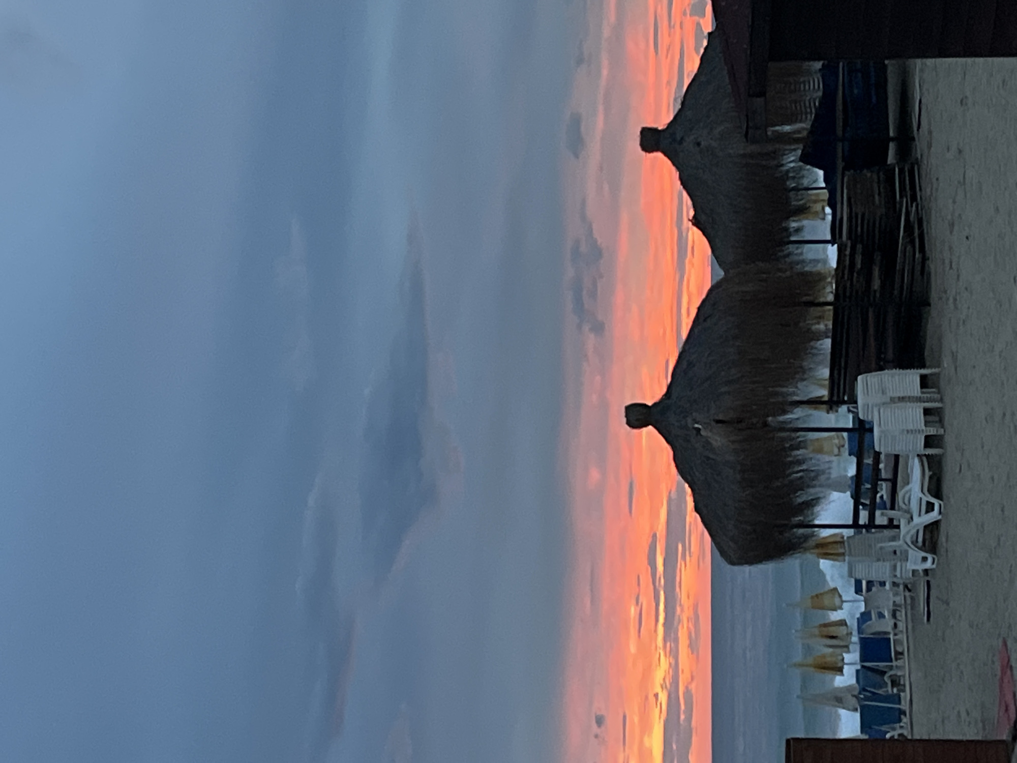The image is of a beach scene in Alanya, Turkey, at either sunrise or sunset. The sky displays hues of orange and pink. Two thatched umbrellas stand on the beach, surrounded by white plastic chairs. The sea is visible in the background, reflecting the colors of the sky. The sand is light-colored. The scene appears devoid of people.