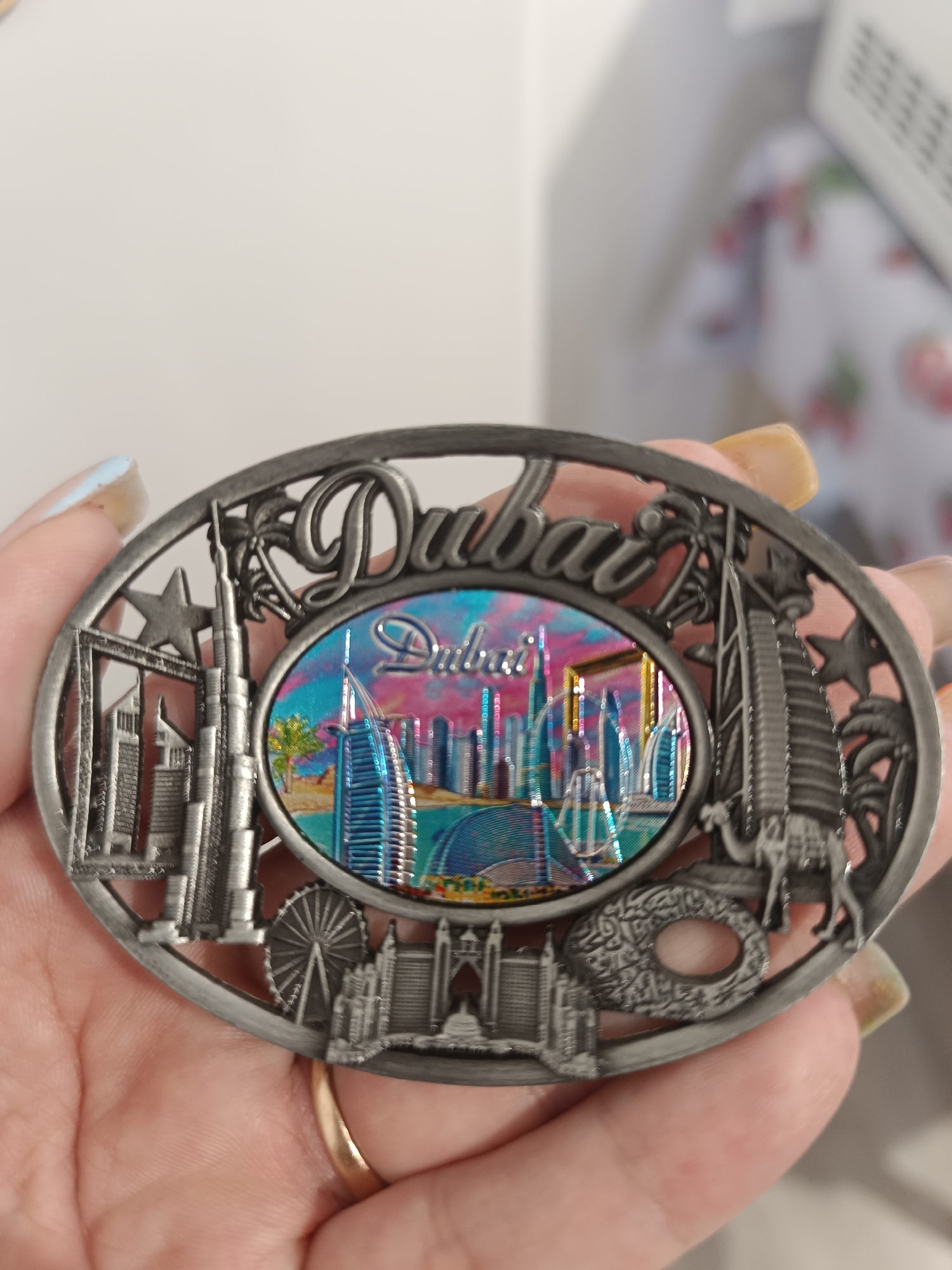 A close-up shot shows a person's hand holding a metal souvenir of Dubai. The souvenir is oval-shaped and features an intricate metal relief of Dubai's iconic landmarks, including skyscrapers like the Burj Khalifa and the Burj Al Arab, a Ferris wheel, and palm trees. The center of the souvenir displays a colorful, iridescent image of the Dubai skyline. The hand is visible from the knuckles to the fingertips, with painted fingernails. The background is blurred, suggesting an indoor setting. The souvenir does not have any association with Taraz city administration, Kazakhstan.