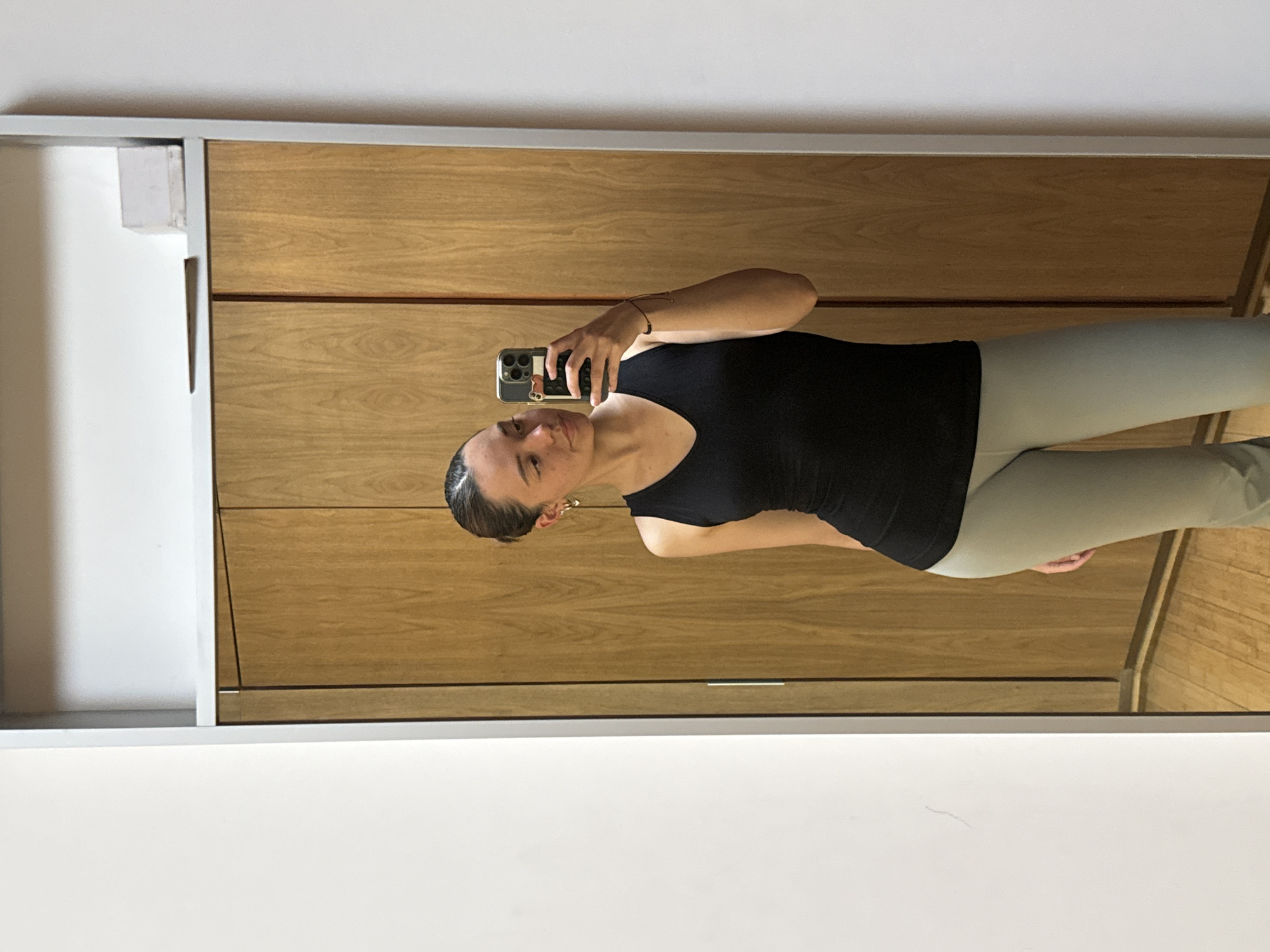 The image depicts an indoor scene, primarily captured through a mirror. No discernible features within the image indicate a specific geographic location such as San Bernardino Tlaxcalancingo, Mexico.
A young adult female, with light skin and dark hair pulled back, is reflected in a large, rectangular mirror framed by a light-colored border. She is wearing a black V-neck tank top and light-colored (possibly light grey or pale green) fitted pants. Her left arm is raised, holding a dark-colored smartphone with a light-colored case featuring a small red heart detail, angled to capture her reflection. She wears small gold-colored hoop earrings, one with a heart charm, and a dark-colored string bracelet on her right wrist. Her gaze is directed towards the smartphone screen.
The background visible in the mirror's reflection consists primarily of two vertical wooden panels, likely part of a wardrobe or closet, with light-colored horizontal handles. Above these panels, a white shelf is visible, upon which a small, clear, rectangular object and a thin, dark, horizontal object rest. To the immediate left of the wooden panels, a white wall is visible. The bottom portion of the reflection shows a wooden floor. The visible area outside the mirror's frame on the left consists of a plain white wall.