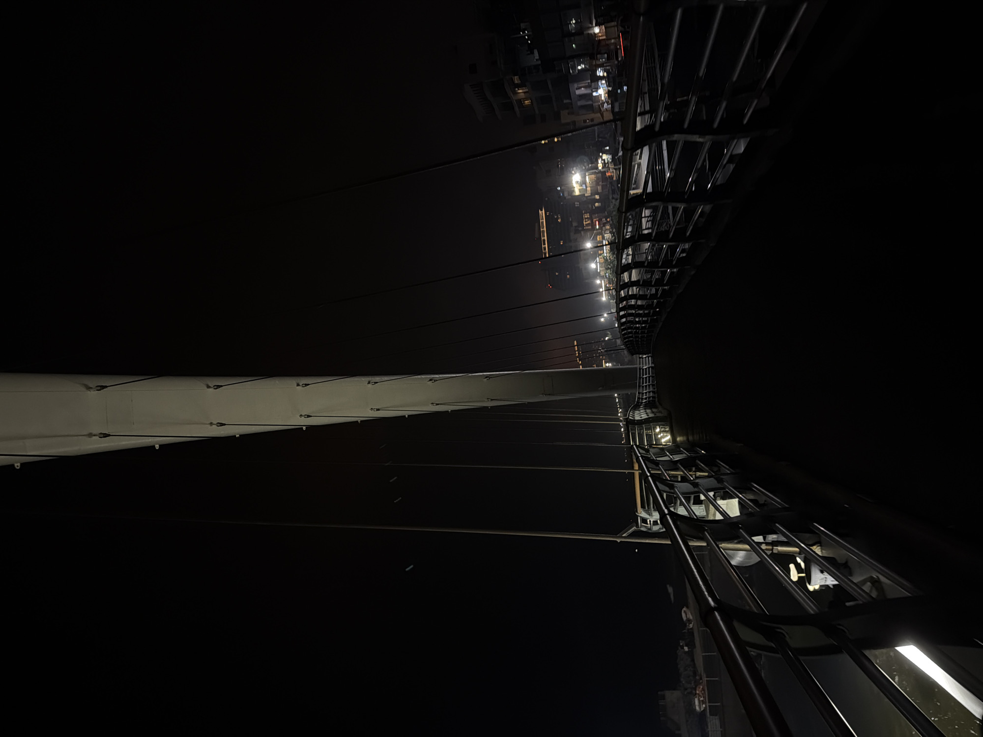The image shows a pedestrian bridge at night in Gangneung-si, South Korea. The bridge is lit by overhead lights and lights from buildings in the distance. The bridge is made of metal and has a modern design with a large white pylon supporting cables. The walkway of the bridge is dark and casts long shadows. There are no people visible in the image. The environment appears to be urban with buildings and a body of water visible.