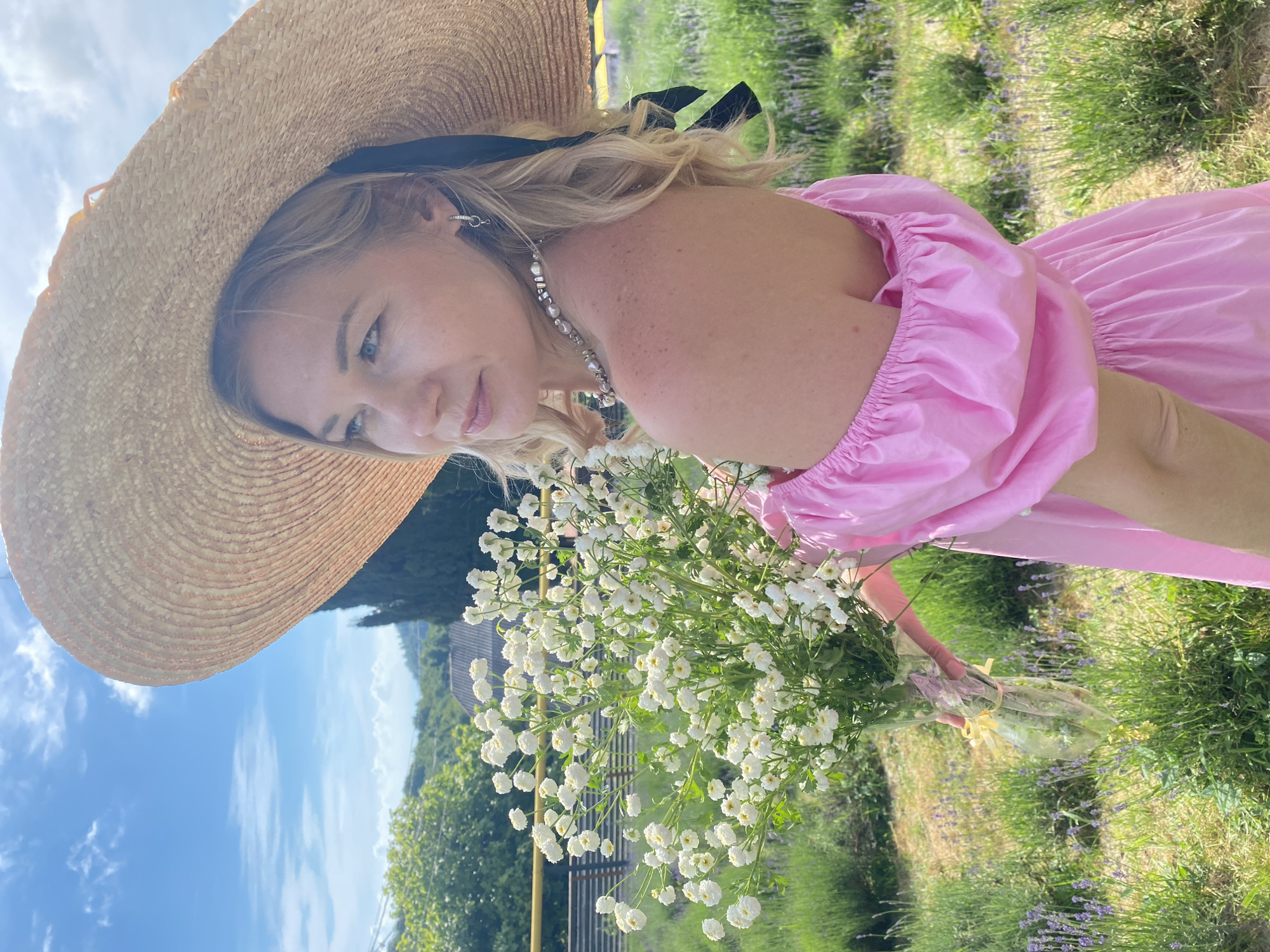 A blonde woman stands in a field of lavender wearing a wide-brimmed straw hat and a pink off-the-shoulder dress. She holds a bouquet of small white flowers. The background features rolling green hills and a clear blue sky. The woman is looking to her left, her expression serene. The scene appears to be outdoors, likely in a rural or garden setting. The location is identified as Verkhneye Uchdere, Russia.