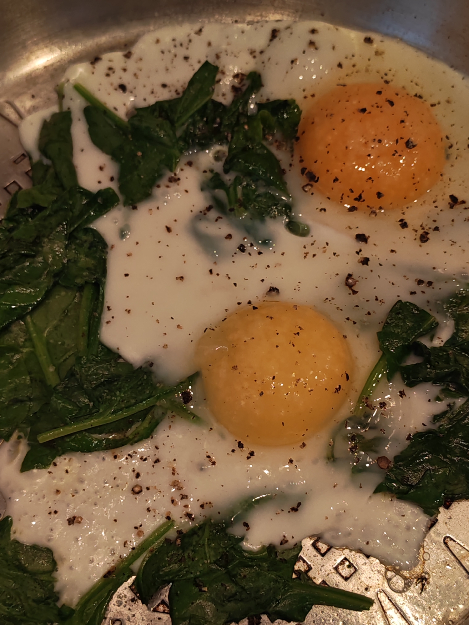 The image shows two fried eggs with spinach and cracked black pepper in a metal pan. The eggs are sunny-side up, with visible yolks. The spinach is wilted and mixed with the egg whites. The pan appears to be on a stovetop, and there is some condensation or steam visible around the food. The lighting suggests an indoor setting, likely a kitchen. There are no people visible in the image. The specific location of Bataysk, Russia, cannot be determined from the visual information provided in the image.