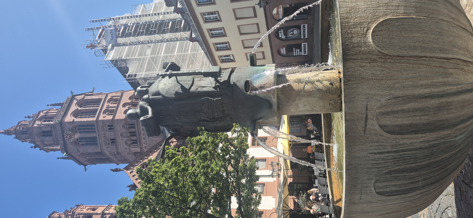 The image captures a public fountain in Neustadt, Mainz, Germany, with the Mainz Cathedral prominently in the background. The fountain features a dark, sculptural element at its center, from which water flows into a large, rough-hewn basin. Behind the fountain, a vibrant green tree partially obscures the ornate facade of the Mainz Cathedral, a Romanesque cathedral with tall spires and clock faces. To the right of the cathedral, a building is covered in scaffolding and white netting, indicating ongoing construction or renovation. In the middle ground, a cluster of outdoor cafe tables and chairs are set up, with several patrons seated and enjoying the sunny weather. The area is paved with cobblestones. Storefronts are visible in the background, one with signage for "MEN'S HAIRCUT" and "GUUDE MEENZER," suggesting a commercial district. The sky is clear and blue.