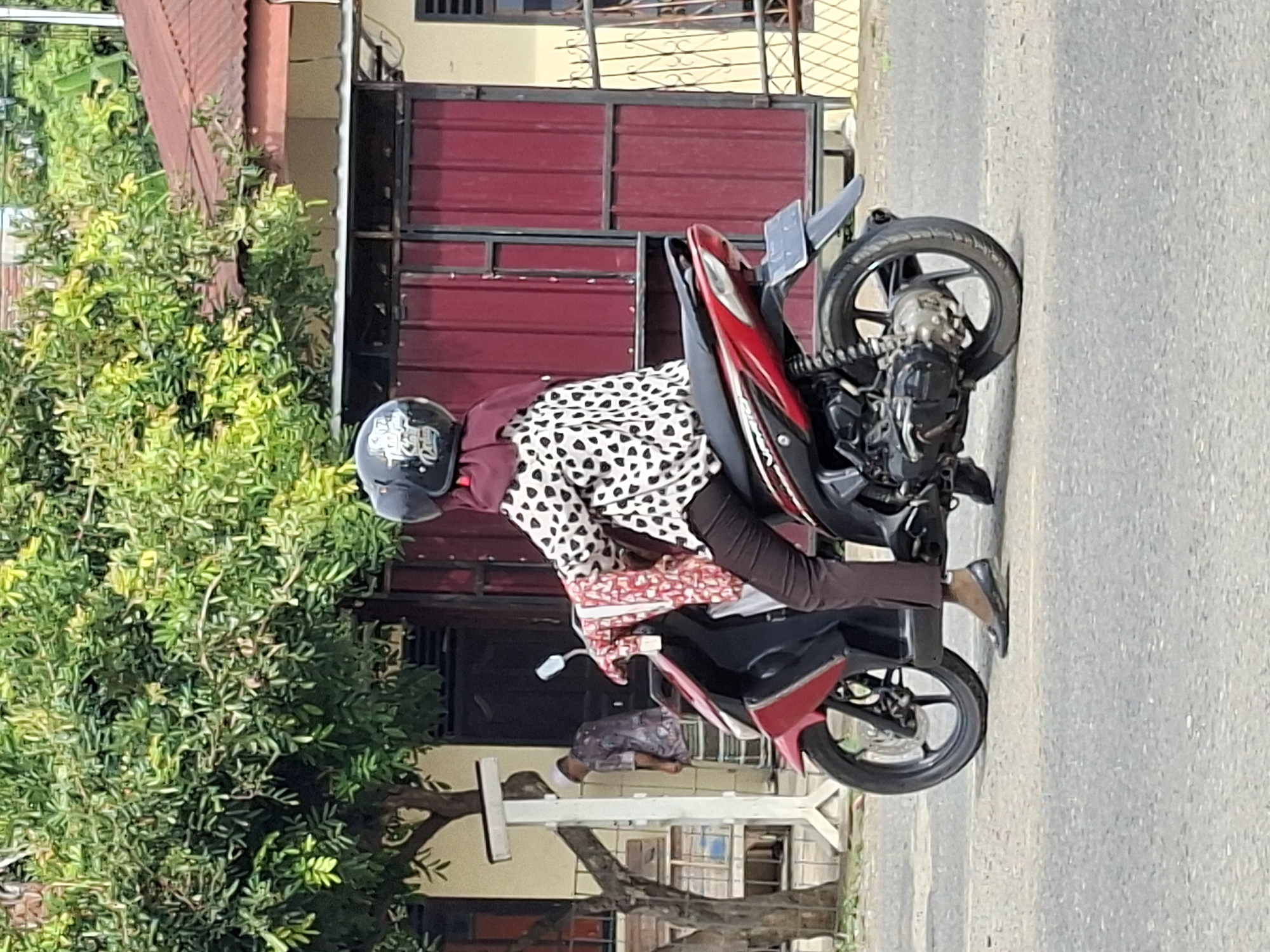 A woman wearing a maroon hijab and a white shirt with black polka dots is sitting on a red and black scooter. She is wearing dark brown pants and black flat shoes. A red and white patterned fabric is draped over her lap. The scooter has a black helmet on the handlebar. In the background, there is a person wearing a patterned shirt and a white cap standing near a white pole with a sign. Behind them is a building with a red metal door and a brown roof, surrounded by green trees. The scene appears to be on a paved street. The location is identified as Lhokseumawe, Indonesia.