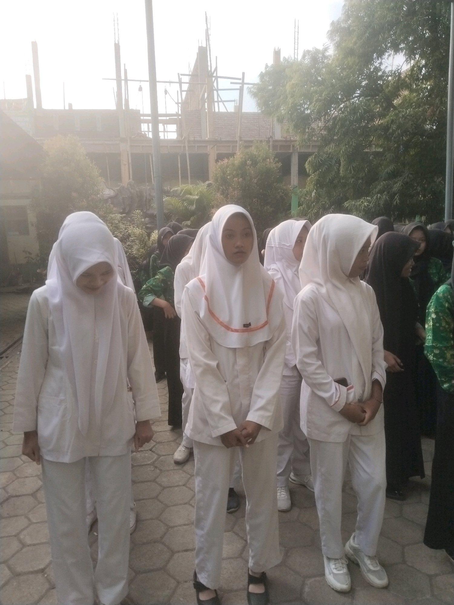 In the city of Pakulaut, Indonesia, several young women, appearing to be students, stand outdoors on a paved walkway. They are wearing matching white uniforms, consisting of long-sleeved tops and pants, with some girls also wearing white headscarves. One girl in the foreground is looking down. Behind them, a building under construction is visible, with scaffolding and exposed structural elements. Trees and foliage are present in the background, suggesting an outdoor campus or public area. The lighting indicates it is daytime, possibly late afternoon due to a slight haze or overexposure in the sky. The overall scene appears to be a casual gathering or possibly a school event.