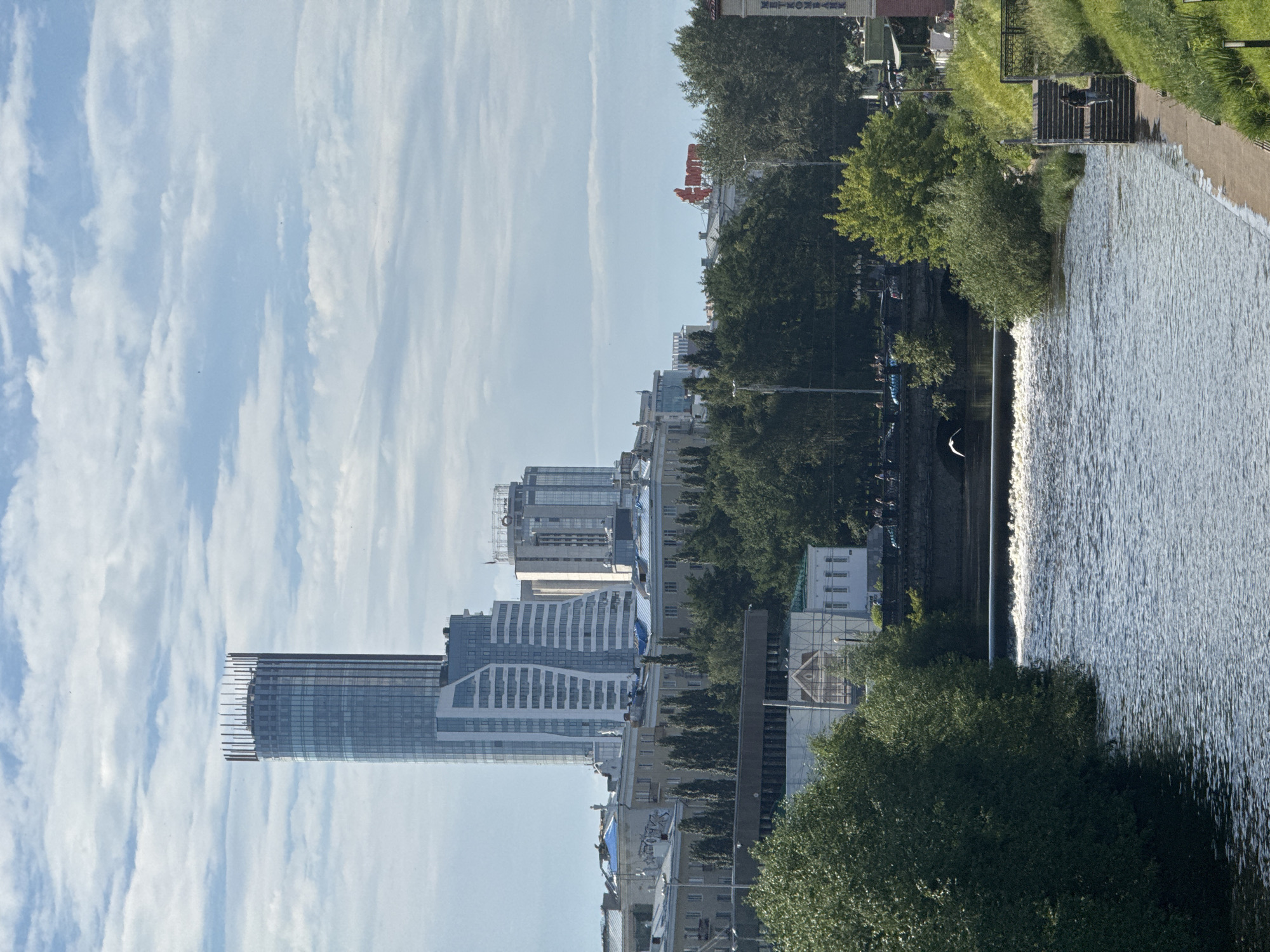The image depicts a waterfront urban scene, likely in Yekaterinburg, Russia. A wide, choppy body of water dominates the right side of the frame, with a visible weir creating a small waterfall. On the left bank of the water, a walkway or path runs alongside a grassy embankment. Further inland, a cityscape rises, characterized by a mix of modern and older architecture. Several tall buildings, including a prominent glass-fronted skyscraper under construction with exposed structural elements, form the skyline. Amidst the buildings, dense green trees line the waterfront, interspersed with smaller structures and what appear to be streets with some vehicular traffic. A few people are visible: one individual stands on a platform overlooking the water, and others are subtly present in the background, suggesting a public space. The sky is a light blue with wispy, elongated clouds, indicating fair weather, possibly late morning or early afternoon. The overall impression is one of a revitalized urban area with a focus on waterfront development. No discernible text is visible within the image.