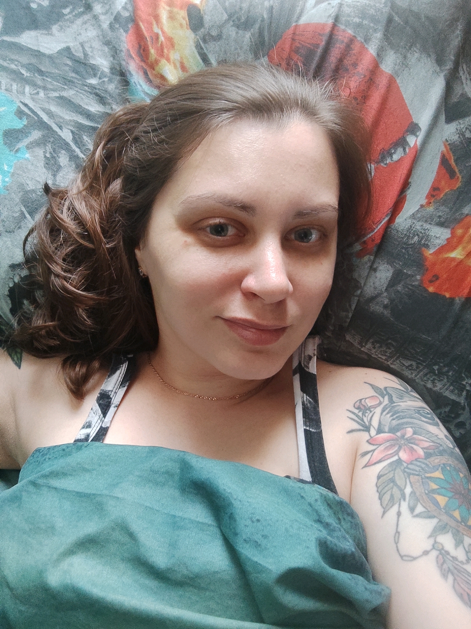 A medium-shot, overhead, indoor portrait shows a woman with medium brown, curly hair lying on a patterned blanket. The woman is facing the camera and looking to her right. She has blue eyes and is wearing a dark-colored camisole with a black and white geometric pattern. A teal-colored cloth is draped over her chest and left shoulder. Her right arm, which is tattooed, is visible on the right side of the frame. The blanket behind her has a repeating abstract pattern of red and orange shapes on a dark gray background. The lighting appears to be natural, coming from above.
