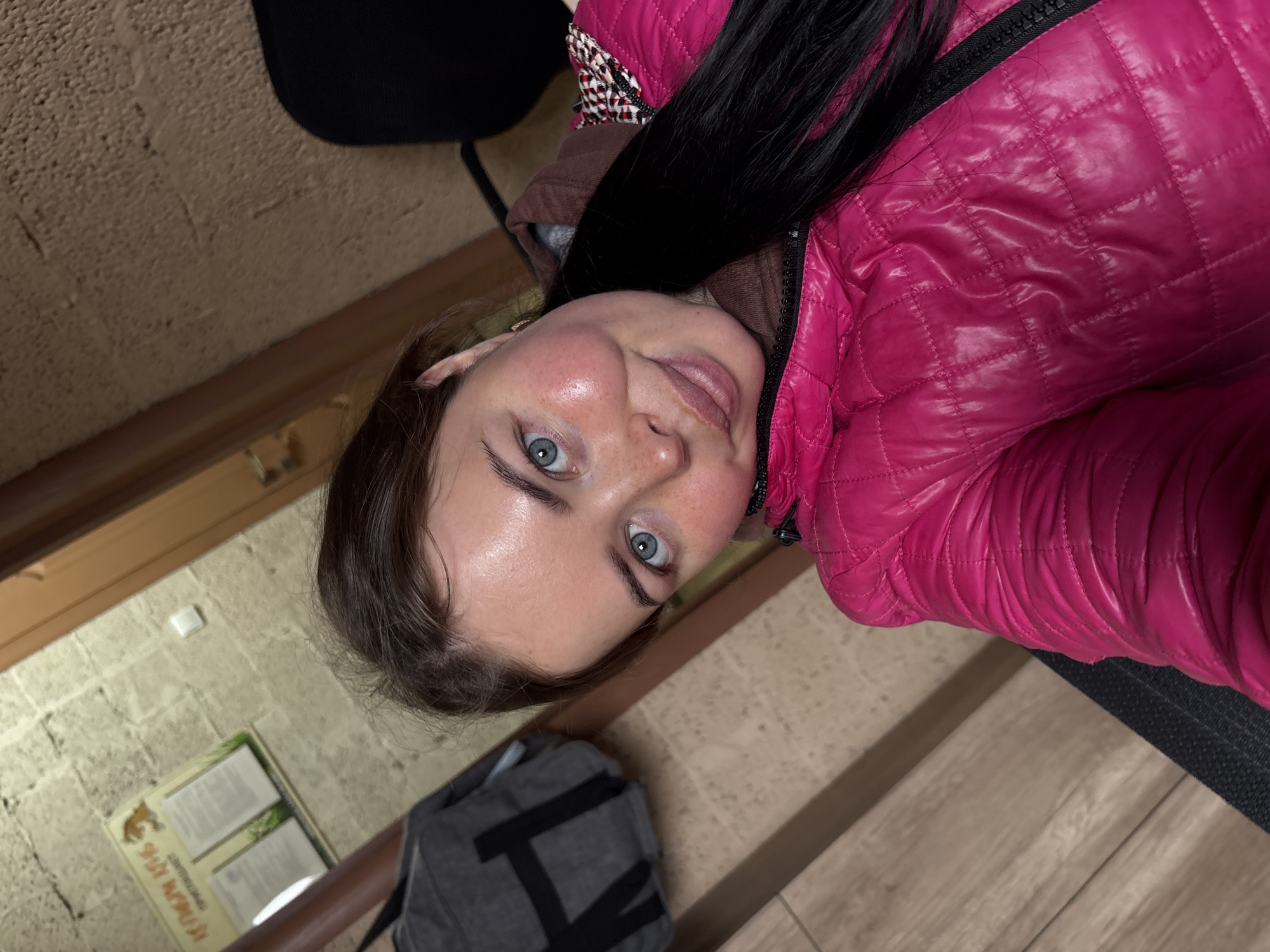 The image is a close-up selfie of a young woman, taken indoors. She has fair skin, light blue eyes, and long dark hair, and is looking directly at the camera with a slight smile. She is wearing a bright fuchsia pink quilted puffer jacket with a black zipper, and a dark-colored, possibly brown, hooded garment underneath. A small, light-colored earring is visible on her right ear.
The background shows an interior setting with varied textures. To her right, a light brown, textured wall, possibly stone or rough plaster, is visible. To her left, a light wooden floor with visible planks is present, on which a dark grey backpack with a black 'V' logo rests. A dark brown wooden trim or railing runs horizontally across the mid-ground.
Further in the background, on the textured wall, a white square object (likely a light switch) is visible above a blurry display board. Partially discernible Cyrillic text on the board includes "РУССКИЙ ВОЖОЙ" and "ЗАПОВЕДНЫЙ КРАЙ", indicating the location is likely a protected area in Русский Вожой, Russia. The scene conveys a casual and relaxed atmosphere, possibly in a visitor center or public building.