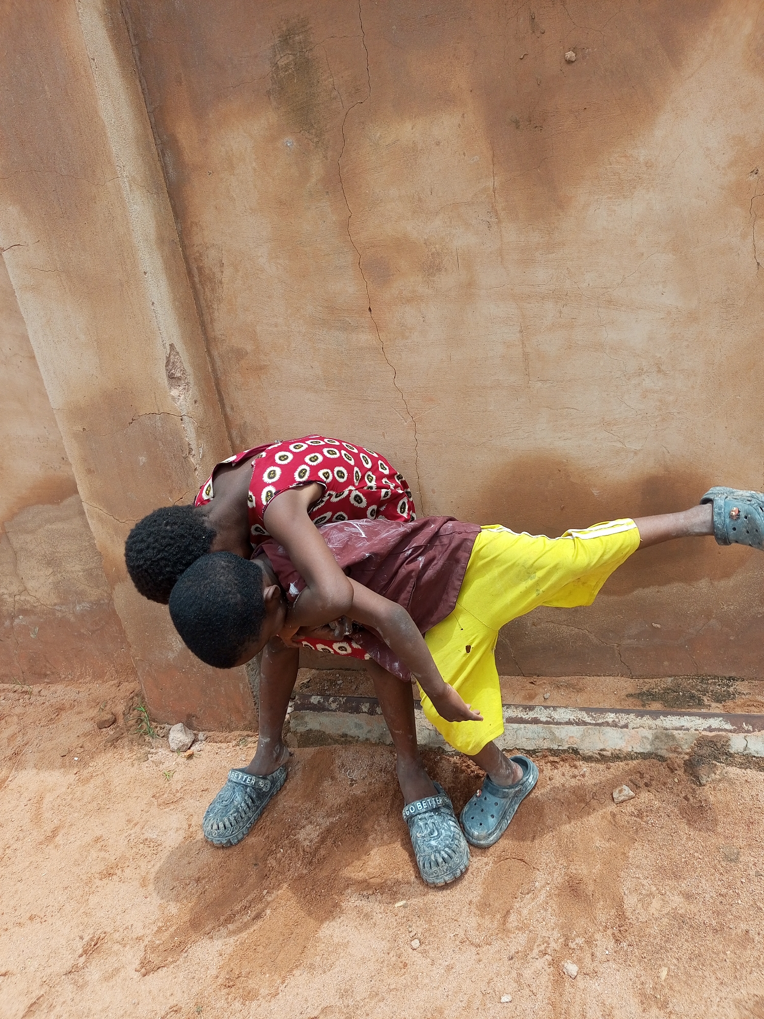 Two children are interacting in front of a textured, ochre-colored wall. The child on the right is positioned horizontally, appearing to be held or supported by the child on the left. The child on the right wears yellow shorts and a maroon shirt and has their right leg extended, wearing a blue clog-style shoe. The child on the left wears a red and white patterned dress and is bent over, with their head near the other child's. Both children have dark, curly hair and are wearing similar blue clog-style shoes. The ground is composed of light brown sand, and there is some debris and sparse vegetation visible. The wall shows signs of age, with cracks and variations in color. The scene appears to be outdoors in an informal setting. It is not possible to determine the location within Tempure, Nigeria from the image.