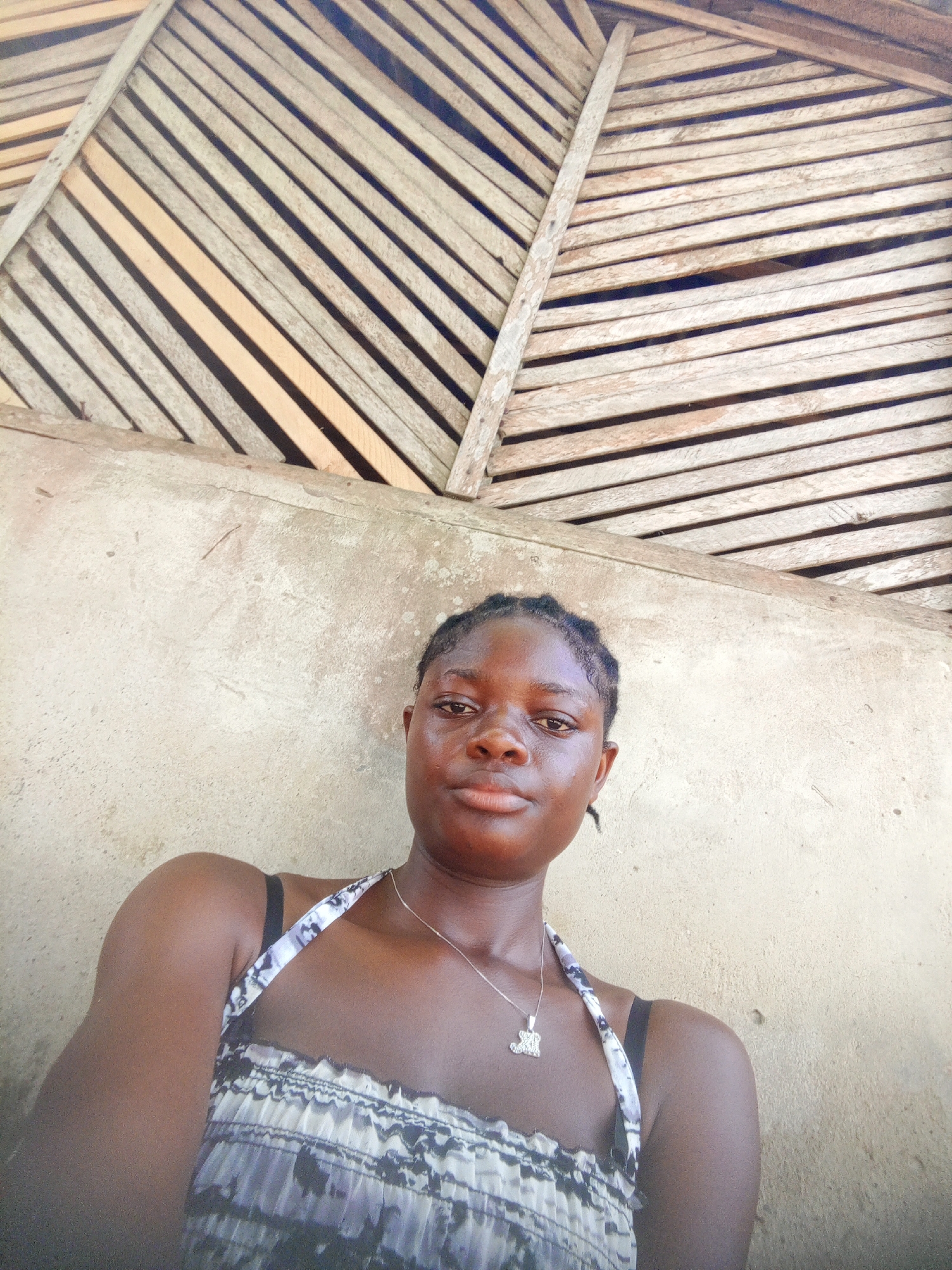 A young woman is the subject of a selfie. She has dark skin and her hair is styled in braids. She is wearing a patterned sleeveless top with thin straps and a silver necklace with a pendant. She is positioned in front of a roughly textured, light-colored wall. Above the wall is a ceiling constructed of horizontally arranged wooden slats. The photograph appears to be taken outdoors, possibly in Mbribit, Nigeria, based on the prompt. The scene seems to be a candid, unposed shot.