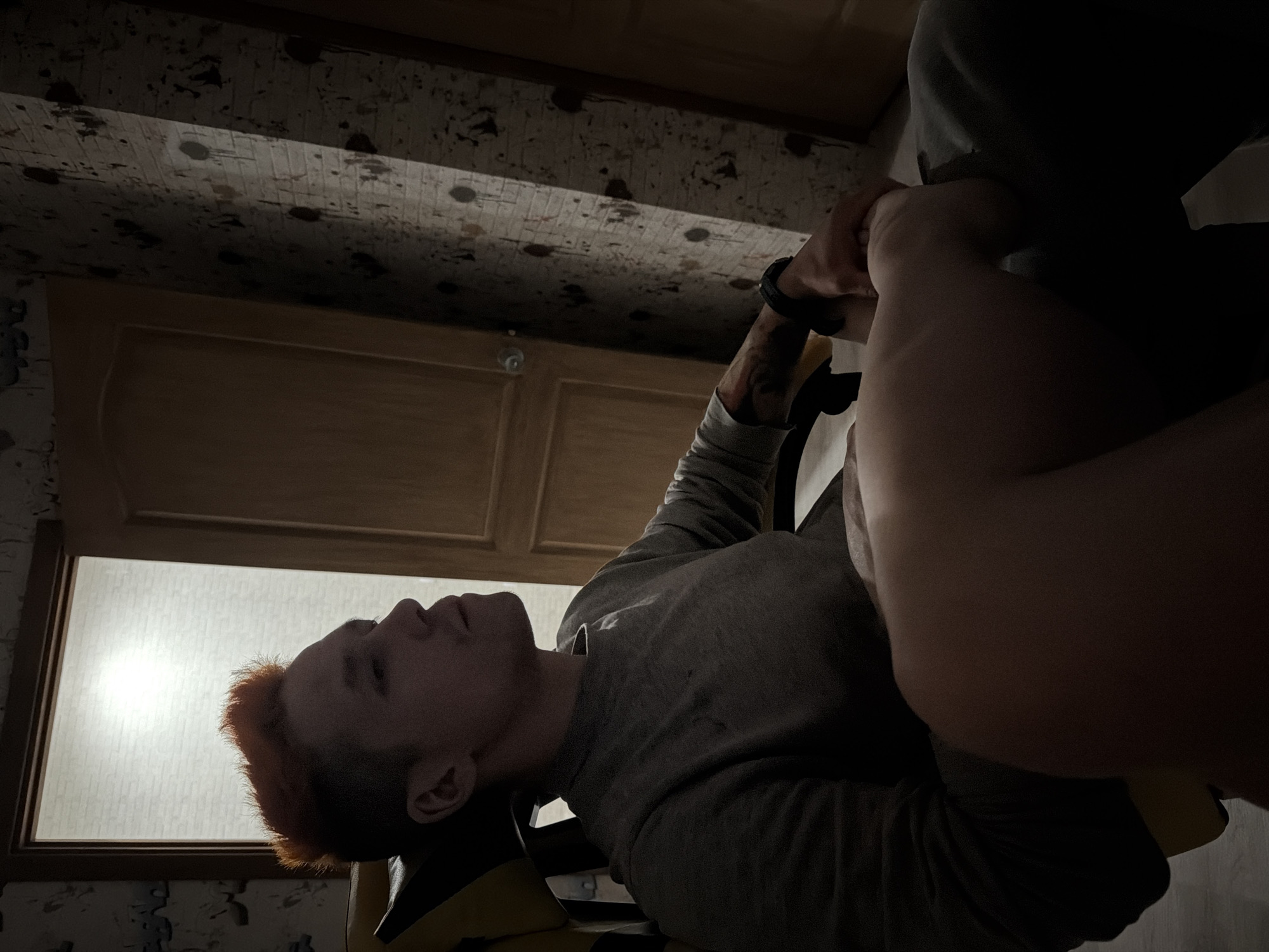 A person with short, reddish hair, wearing a gray long-sleeved shirt, is reclined in a chair within an interior space. The chair has yellow and black elements. The person's left wrist has a dark-colored watch or bracelet. A second individual or part of one is significantly closer to the camera, obscuring part of the view. A window is visible in the background, providing indirect illumination. Above the person, there is a wooden door and patterned wallpaper. The location is indicated as Chaikovsky, Russia.