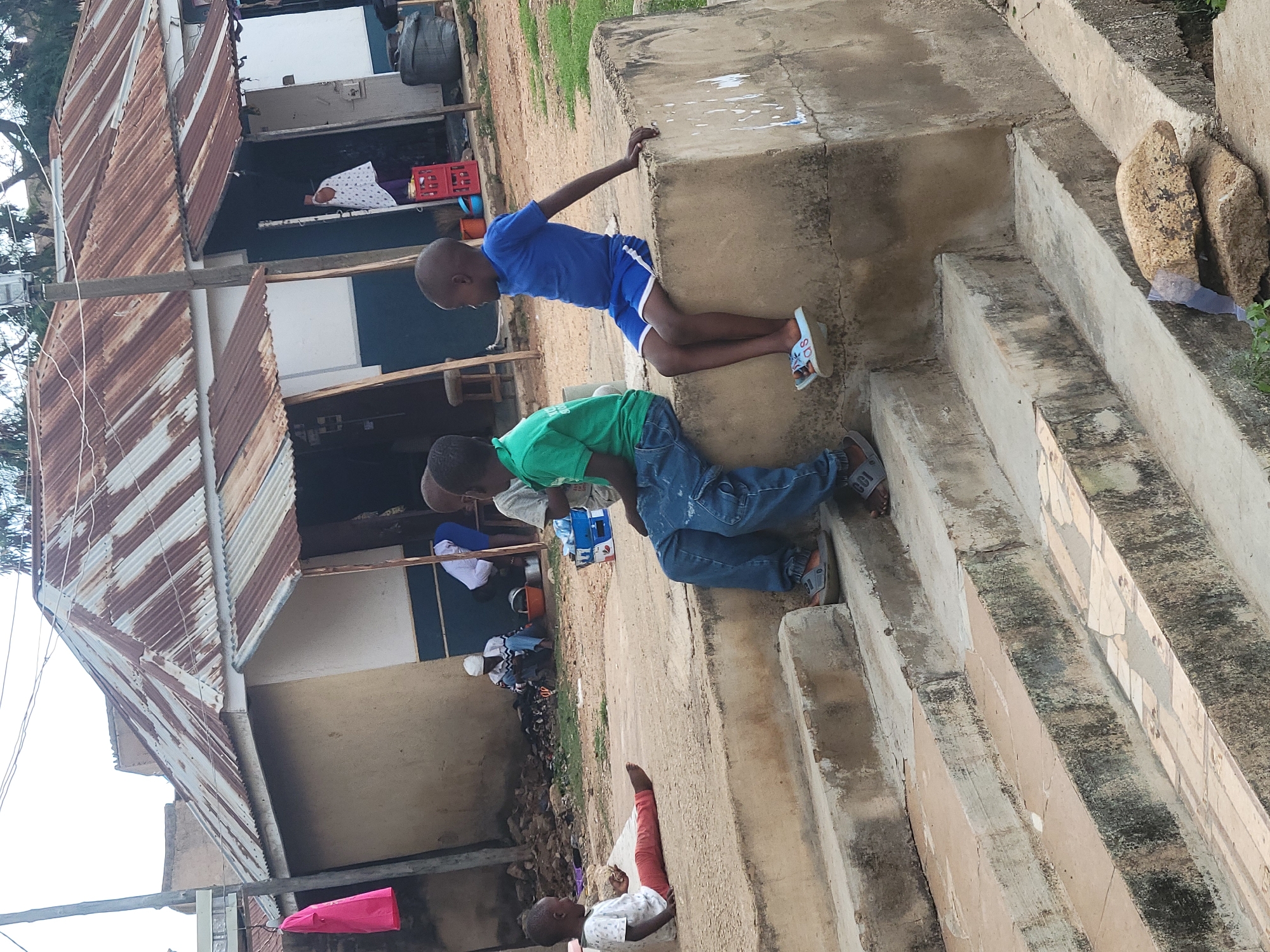 The image shows a scene in Jos, Nigeria, with a building on the left featuring a corrugated iron roof. On the right are concrete steps leading to an elevated platform. Two boys are positioned on the steps and platform. One wears a blue shirt and shorts, and rests on the platform. The other wears a green shirt and jeans, and is positioned on the steps below. In the background, several people can be seen near the building.