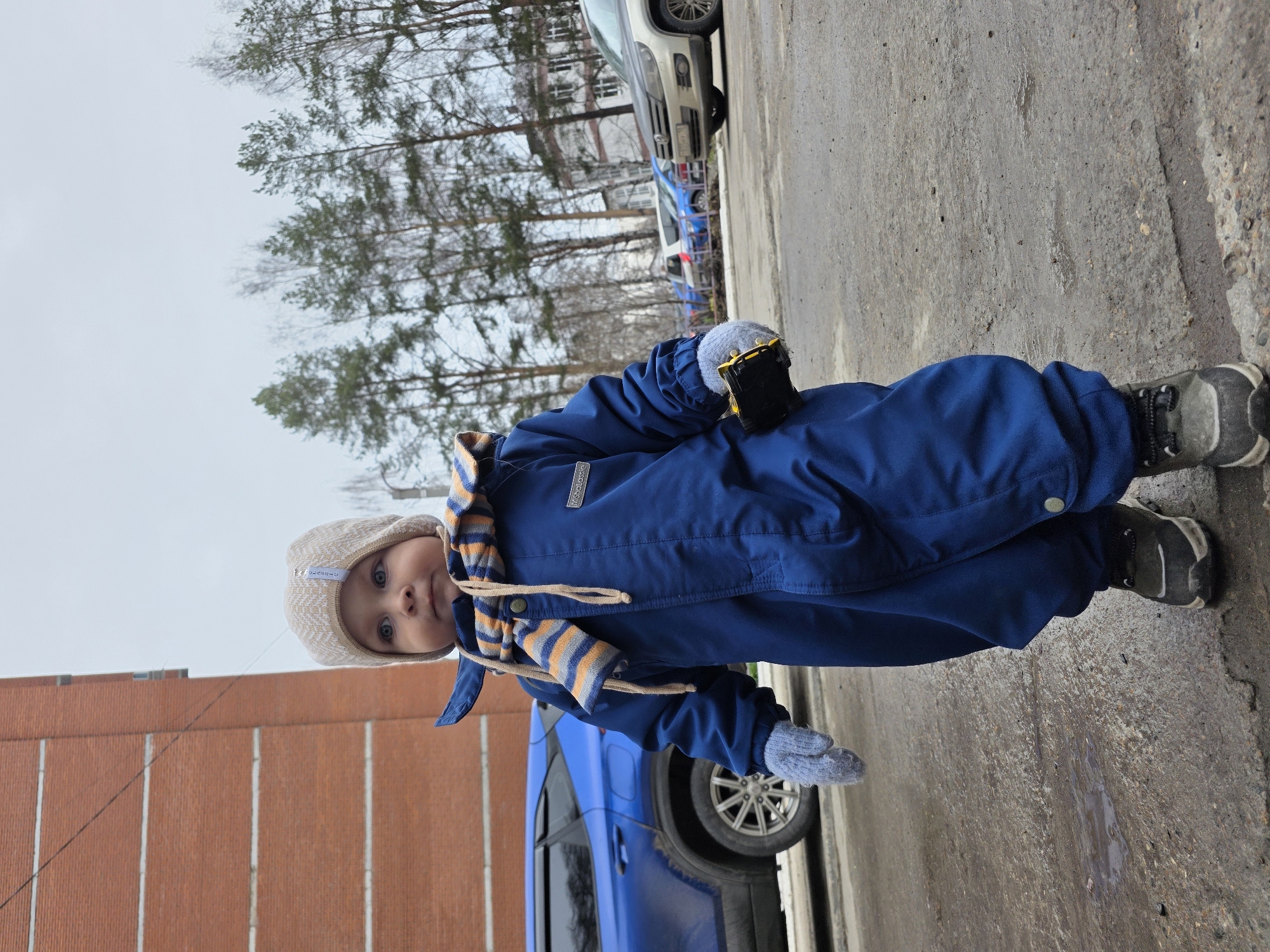 A young child, wearing a blue snowsuit, a beige hat, and striped scarf, is standing on a paved surface. The child is also wearing grey mittens and dark-colored shoes with grey soles. In the child's gloved right hand, they hold a small, dark-colored toy with yellow accents. Behind the child, there's a brick building on the left, a blue car, and a silver car. In the background, there are trees, and possibly other buildings partially visible. The scene appears to be outdoors, possibly in a residential area in Chaikovsky, Russia.