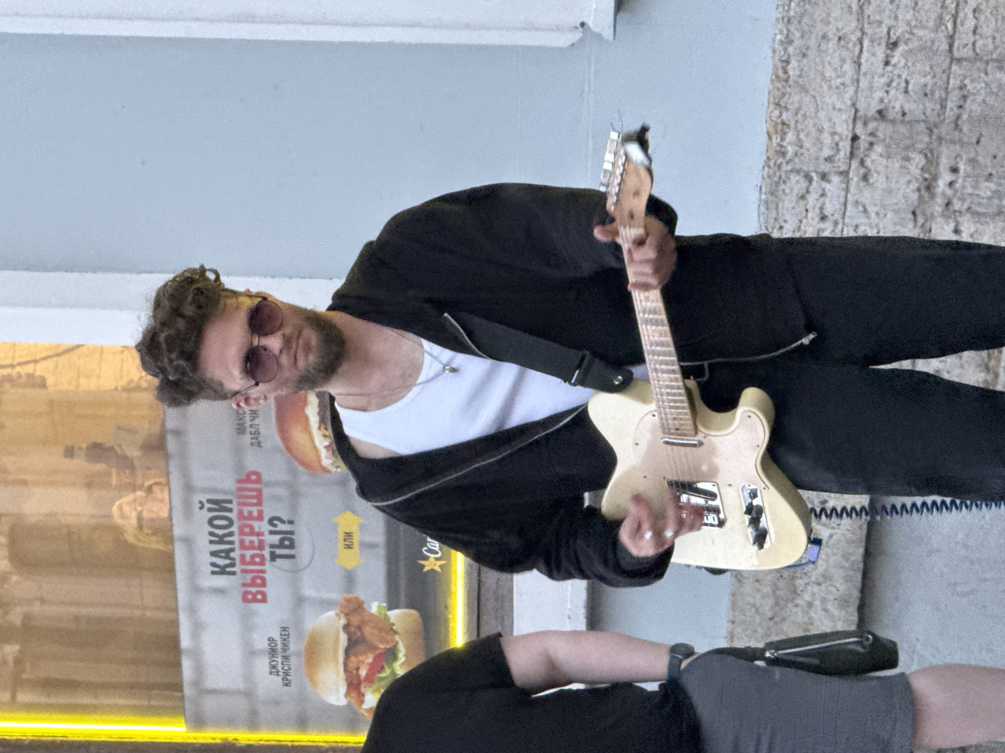 A young man with curly brown hair and sunglasses is playing a light-colored electric guitar in front of a brick wall. He is wearing a white t-shirt, a black zip-up hoodie, and black pants. A black cable runs from the guitar to the ground. Behind him, a large poster displays images of burgers and text in Russian, including "КАКОЙ ВЫБЕРЕШЬ ТЫ?" (Which will you choose?). The man appears to be standing on a sidewalk, with the building facade and a street visible in the background. The lighting suggests it is daytime.