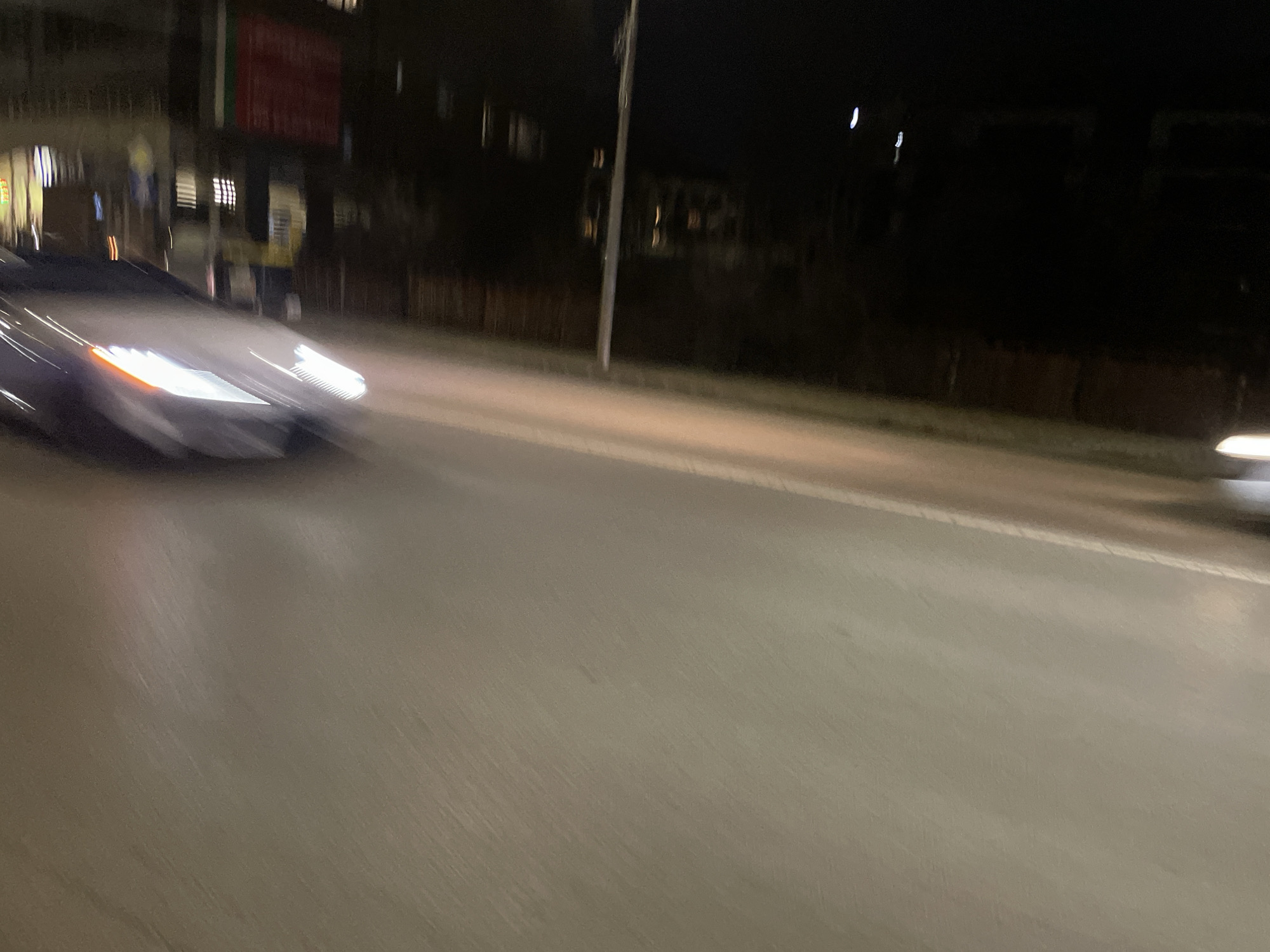 The image depicts a nighttime urban scene, likely a street in Sofia, Bulgaria, based on the visible building styles and street lighting. A car is moving rapidly from left to right, indicated by the motion blur. The car's headlights are illuminated. In the background, there are indistinct buildings, some with lit windows. A street lamp is visible on the side of the road. The road surface appears to be asphalt. Another car with illuminated headlights can be seen in the distance to the right. The overall lighting is dim, with the primary sources being the car headlights, street lamp, and building lights.