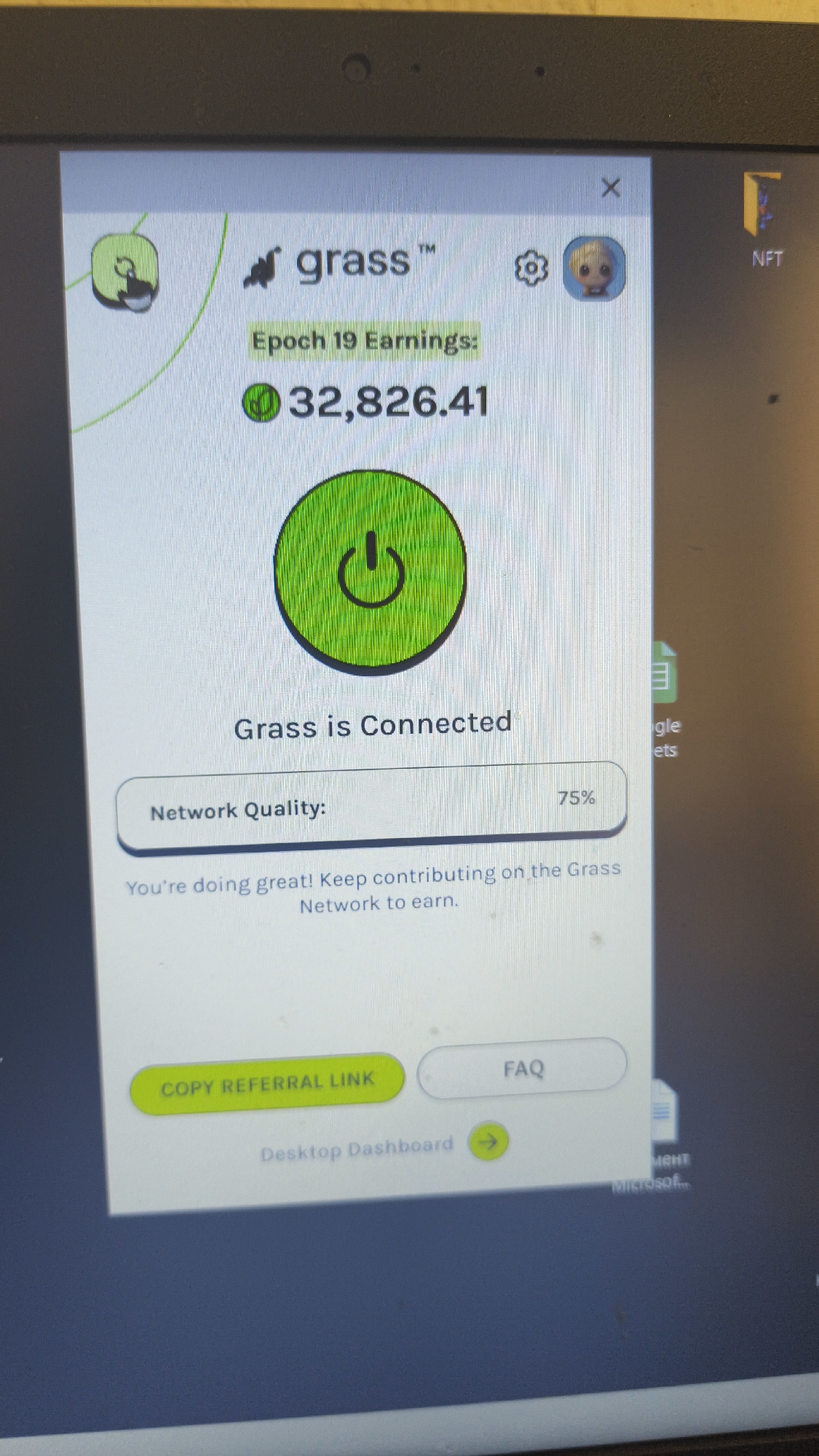 A photograph captures a computer screen displaying an active application called "grass™," likely an extension or pop-up window. The screen's bezel is visible at the top, showing a small circular webcam. The application, primarily in green and white, prominently features "Epoch 19 Earnings: 32,826.41," with a green coin-like icon next to the numerical value. Below this, a large green circular graphic with a power symbol indicates "Grass is Connected."
Further down, a status bar shows "Network Quality: 75%." A message reads, "You're doing great! Keep contributing on the Grass Network to earn." At the bottom of the application window, there are two interactive buttons: a green button labeled "COPY REFERRAL LINK" and a white one labeled "FAQ." Below these, a link "Desktop Dashboard →" is present with a green arrow.
The top bar of the application includes the "grass™" logo, a settings gear icon, a small cartoon avatar, and an "X" button to close the window. The desktop background, partially visible around the application, shows fragments of file icons and text, including "NFT," "gle ets" (likely Google Sheets), and "Microsof" (likely Microsoft). The image is slightly angled, suggesting it was taken by a handheld device.