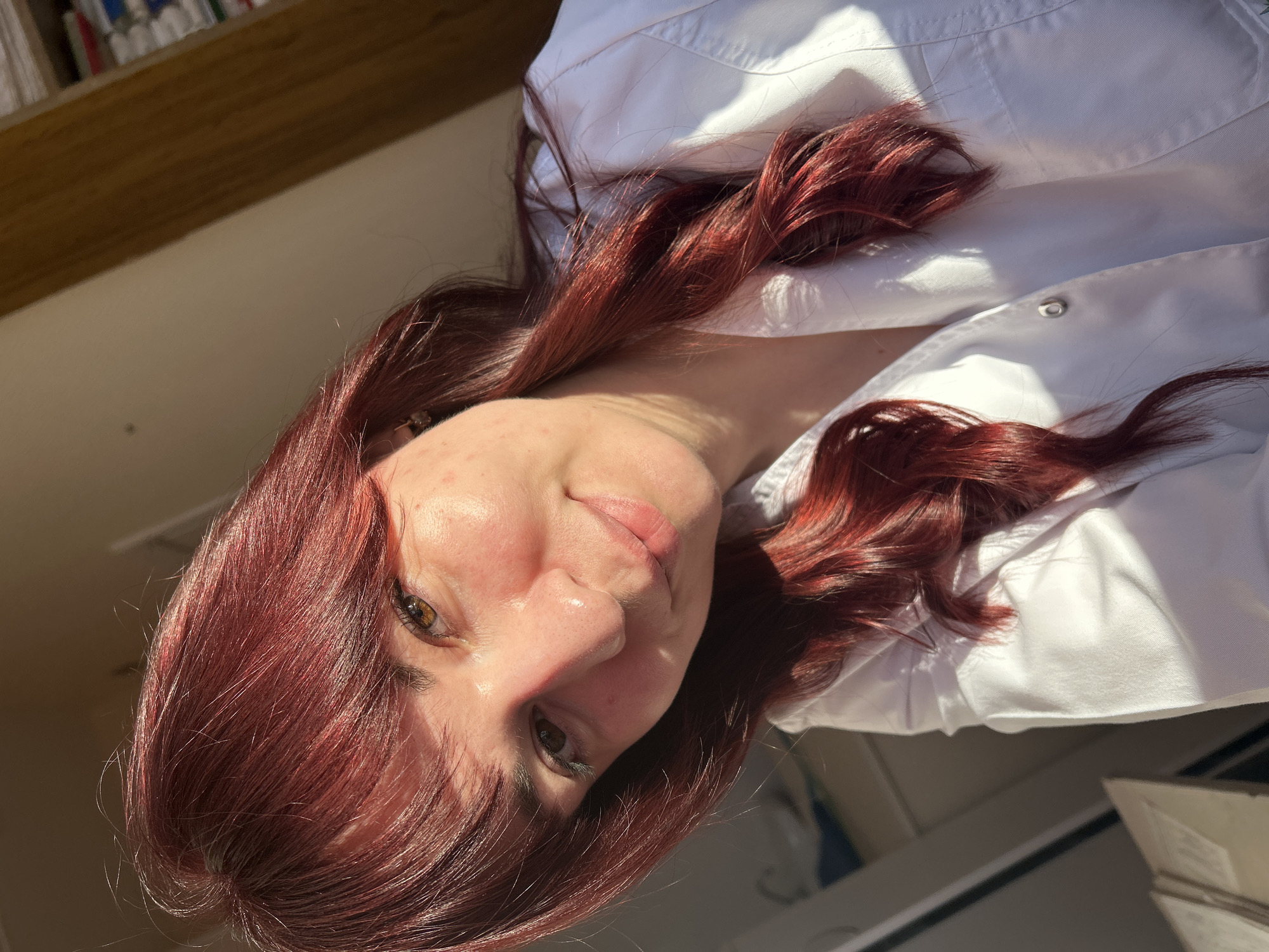 A woman with auburn hair is pictured from a low angle indoors. She is wearing a white button-down shirt. The image appears to be taken near a bookshelf. The location cannot be determined to be in Фоки, Russia.