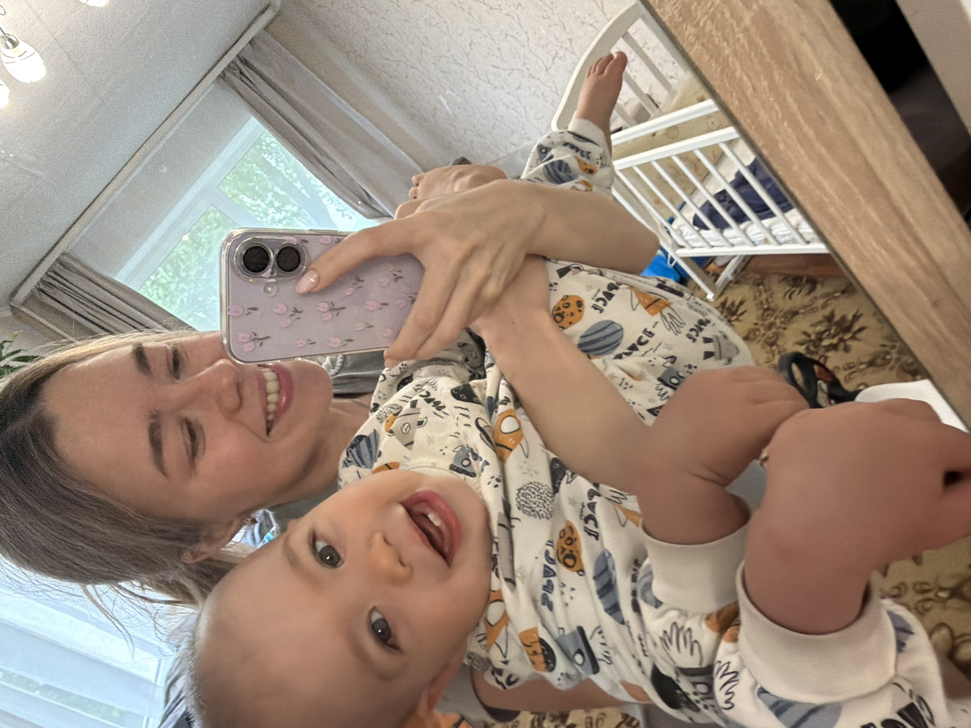 A woman and a baby are in a room in what appears to be an apartment in Chaikovsky, Russia. The woman is holding a phone with a lavender floral case, taking a selfie with the baby. Both are smiling. The baby is wearing a white patterned onesie. In the background is a white crib and a window with curtains. The room also contains a mirror reflecting the scene.