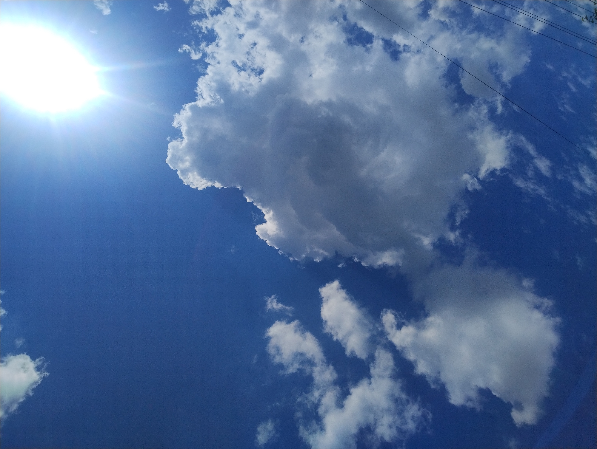 A bright, sun-drenched sky with fluffy white clouds dominates the image, suggesting a clear day. The sun is a powerful, glowing orb in the upper right corner, with visible rays extending outwards. Several clouds are scattered across the deep blue expanse, ranging from small wisps to a large, prominent cloud formation in the center. The scene appears to be an outdoor shot, likely taken from ground level, looking upwards. There are no people or specific city landmarks visible, making it impossible to determine the location within Unknown, Unknown. However, the presence of what appear to be thin, dark lines resembling power lines running diagonally across the lower right portion of the frame hints at the proximity of urban or suburban infrastructure. The overall impression is one of a bright, sunny day with a dynamic sky.