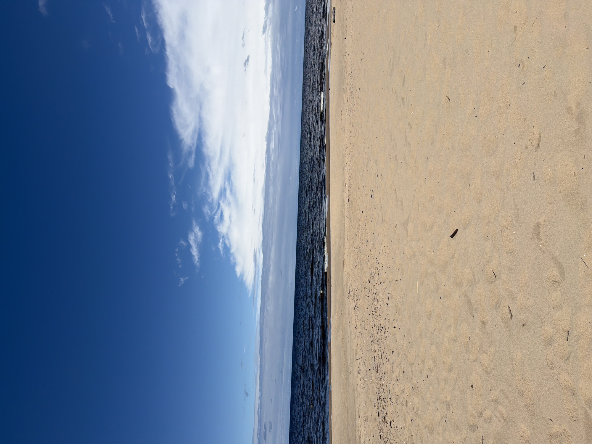 A bright, sunny day at the beach in Lilaste, Latvia, is captured in this vertical, eye-level shot. The scene is divided into three distinct horizontal bands. The top portion of the image is dominated by a vast expanse of clear, deep blue sky, with a few wispy white clouds scattered across it. Below the sky, a dark blue body of water, likely the Baltic Sea, stretches across the frame. The water exhibits gentle ripples, indicative of a light breeze, and the horizon is marked by a thin line of pale clouds. The foreground of the image is a sandy beach, light beige in color, with subtle undulations and scattered small pieces of driftwood and debris. The sand appears dry and undisturbed, suggesting it is not a heavily trafficked area at this moment. There are no people or animals visible in the frame, and the overall impression is one of peaceful solitude and natural beauty.