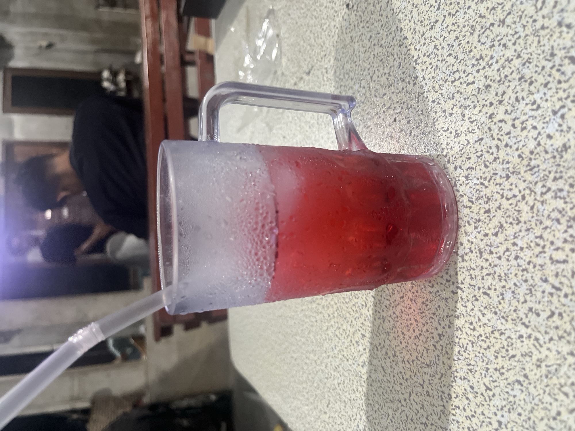 An over-the-shoulder, eye-level shot captures a clear, plastic mug filled with a red beverage and ice, resting on a speckled, light-colored countertop. A white plastic straw extends from the mug at an angle. The mug is condensation-covered. In the blurred background, a wooden bench and a person wearing a black shirt are visible, suggesting an indoor or covered outdoor seating area, possibly a restaurant or cafe. The setting appears to be in Blangseureukuy, Indonesia.