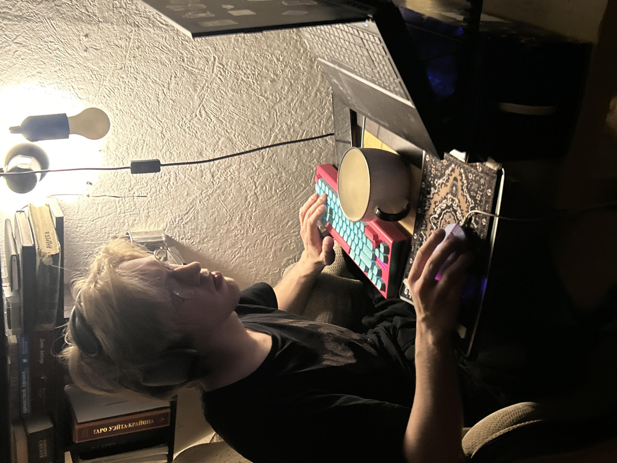 A young person with blonde hair is sitting in a cushioned chair in an indoor setting, using a computer. The person is wearing a black t-shirt and headphones. They are operating a red and teal keyboard with a mug placed on top. A laptop screen is partially visible above the keyboard, and a mouse is positioned to the right on a decorative mousepad.
To the left of the person, a stack of books is visible, with some titles in Russian. Above the person, a lamp with exposed bulbs is attached to the wall. The background is a textured wall.