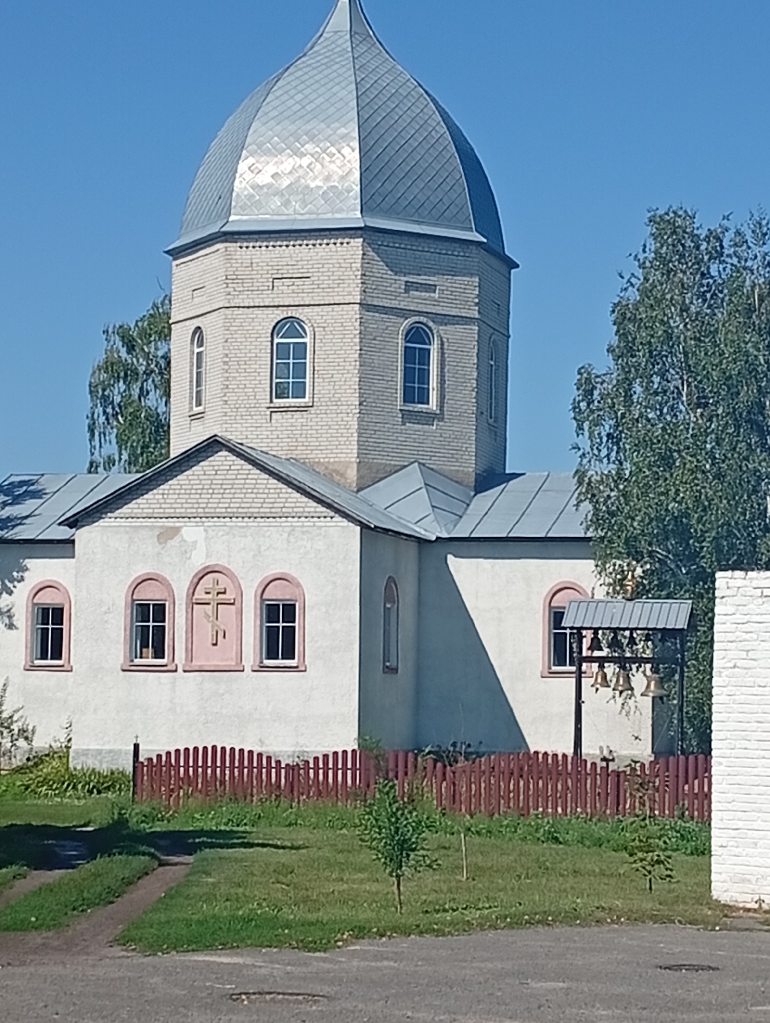 A sunlit, exterior shot captures a light-colored brick church with a grey, faceted dome against a clear blue sky. The church features arched windows, some with light pink trim, and a decorative cross above a central window. A small bell tower with several bells is situated to the right of the main building. A red wooden picket fence runs along the front of the property, separating the church from a grassy area with young trees and a dirt path. The background shows green trees and a portion of a white brick building. The scene is set in Khomutets, Ukraine.