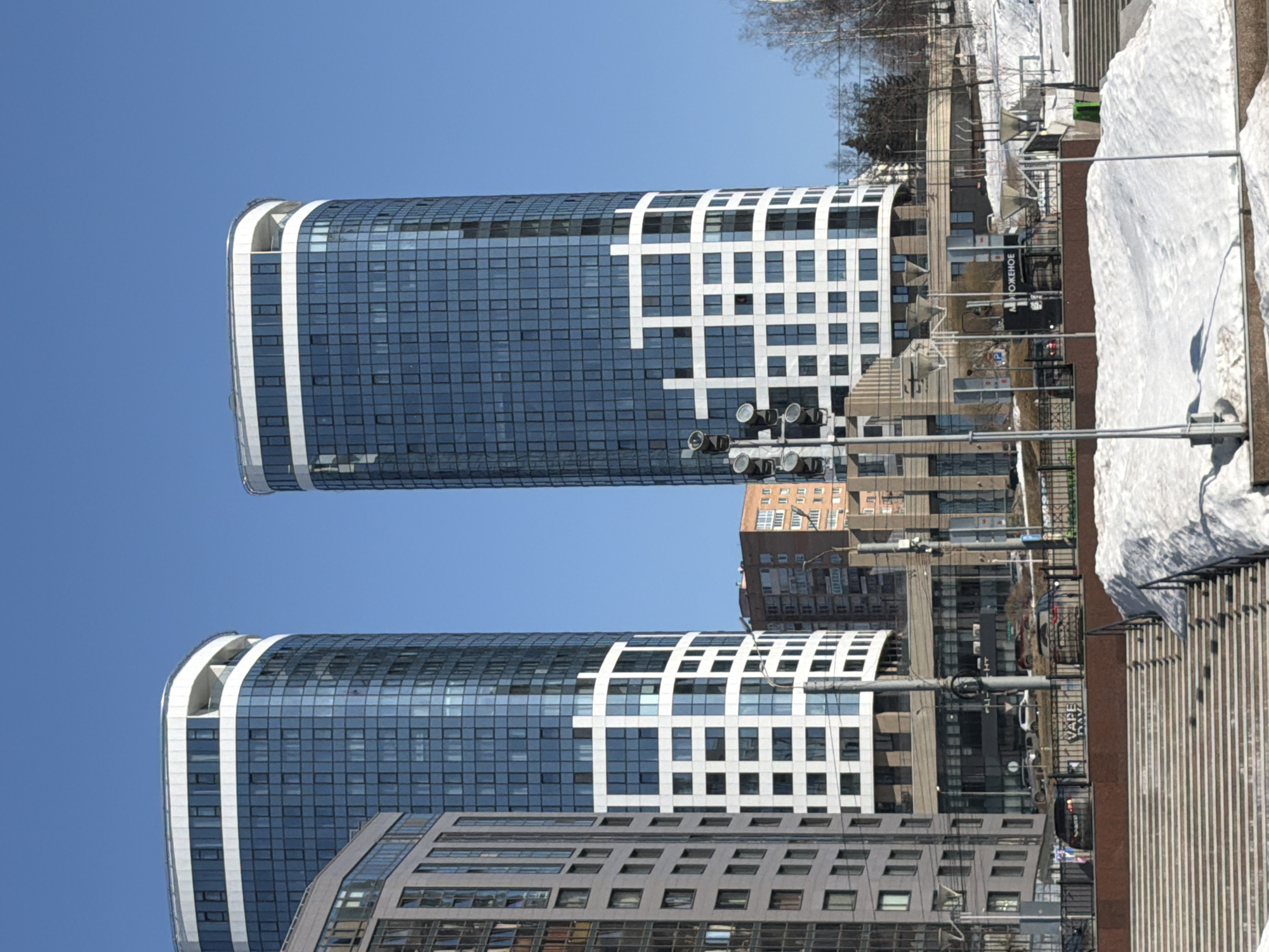 The image depicts a winter or early spring urban scene in Izhevsk, Russia, specifically focusing on the "Bukhara" residential complex located on Pushkinskaya Street. Two prominent, modern high-rise buildings with curved, oval profiles dominate the center-right and center-left of the frame, set against a clear blue sky. Both towers feature dark blue reflective glass facades. The tower on the right exhibits a distinct white grid pattern covering its lower and mid-sections, while both have a horizontal white band and an open top-floor balcony/terrace area.
In the foreground, a wide, multi-tiered concrete staircase with a black metal railing ascends from the lower-left towards the center. Patches of dirty snow are visible on the ground beside the staircase and on an embankment to the right. A tall metal pole with multiple circular traffic signal lights is positioned in the lower-center. Behind the railing, a street or access road is visible with several parked and moving vehicles, including a dark-colored SUV and a light-colored sedan. Signs with Cyrillic text and one partially legible as "VAPE" are visible on structures bordering this road.
Further back, between the two main towers, an older, reddish-brown brick residential building with multiple windows is partially visible. Bare deciduous trees are present on a snow-covered slope in the background-right. No discernible individuals are engaged in specific actions, and the scene is primarily architectural and environmental under clear daylight conditions.