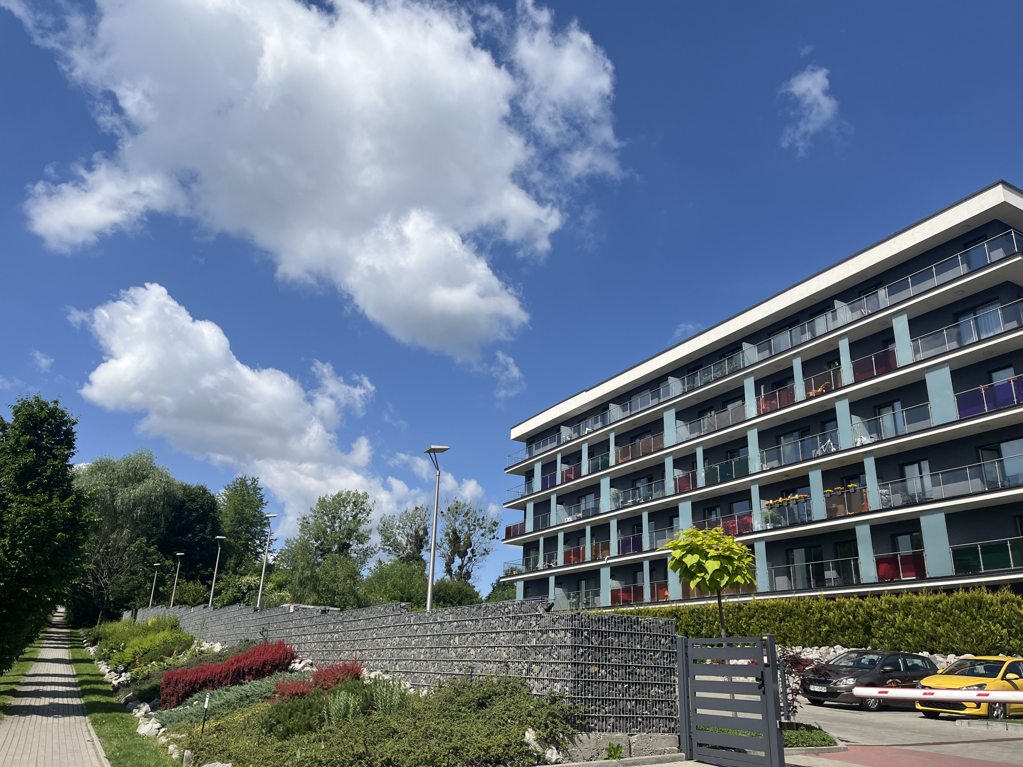 The image shows a modern, multi-story apartment building with a dark blue facade and glass balconies featuring colored panels. In front of the building, there is a well-maintained landscaped area with a gabion wall, a paved walkway, and various types of vegetation including shrubs and flowers. Two cars, a dark brown sedan and a yellow hatchback, are parked in a driveway. The sky is clear and blue with some fluffy white clouds. The scene is outdoors, suggesting a residential area with a contemporary architectural style.