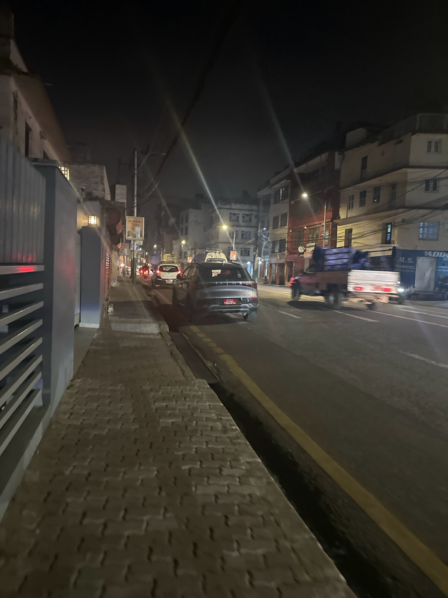 The image depicts a street scene at night in Kathmandu Metropolitan City, Nepal. The perspective is from a pedestrian walkway, looking down a paved street lined with buildings. Several vehicles are present on the street, including a dark-colored SUV in the foreground, a white car further down, and a flatbed truck carrying blue cargo. The street has a yellow dividing line. Buildings on either side of the street are multi-story structures, some with lit windows. Streetlights are visible, casting illumination on the scene. The walkway is constructed of interlocking paving stones. The overall impression is of a moderately busy urban street at night.