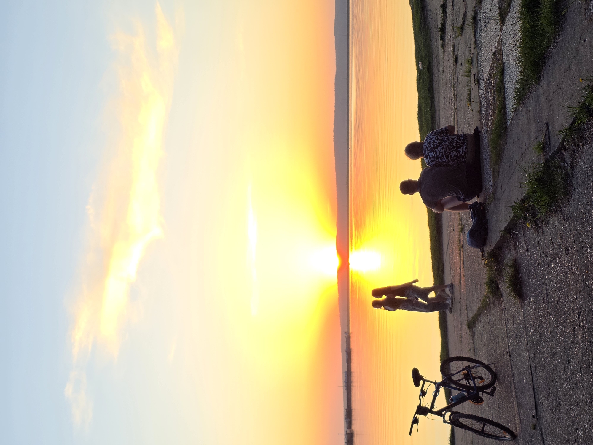 Captured during the evening in the city of Chaikovsky, Russia, the image shows a serene sunset over a large body of water, possibly a lake or reservoir. On the right side of the frame, a concrete embankment is visible, where two people are sitting, and another two are standing nearby, seemingly observing the sunset. A bicycle leans against the embankment. The sky is a blend of light blue and vibrant yellow and orange hues due to the setting sun. The water reflects these colors, enhancing the scene's warm, tranquil atmosphere.