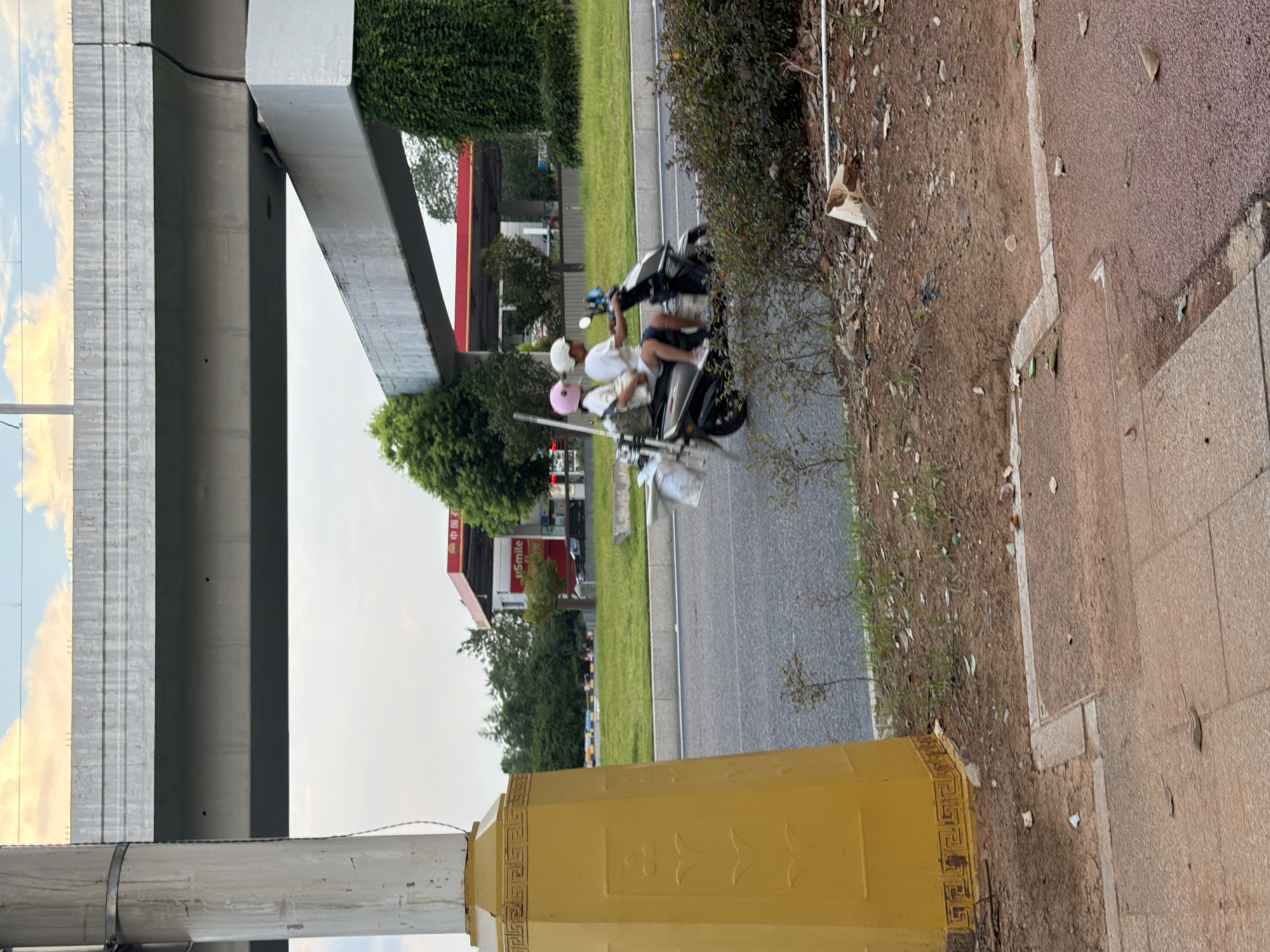 The image shows a street scene in Jiangjin, China, with a two-lane road and an elevated highway overhead. In the foreground, a yellow decorative pillar stands on the left. On the road, a scooter carrying two individuals is moving to the right. The person in the back, wearing a pink helmet, is a female, and the person in the front, wearing a white helmet, is a male. Both are wearing casual clothing. A gas station with the "uSmile" logo is visible in the background, nestled behind trees and lush green foliage. The sky above is partly cloudy with hues of yellow and orange, suggesting it might be late afternoon or early evening. The overall environment appears to be an urban or suburban setting with moderate vegetation and infrastructure.
