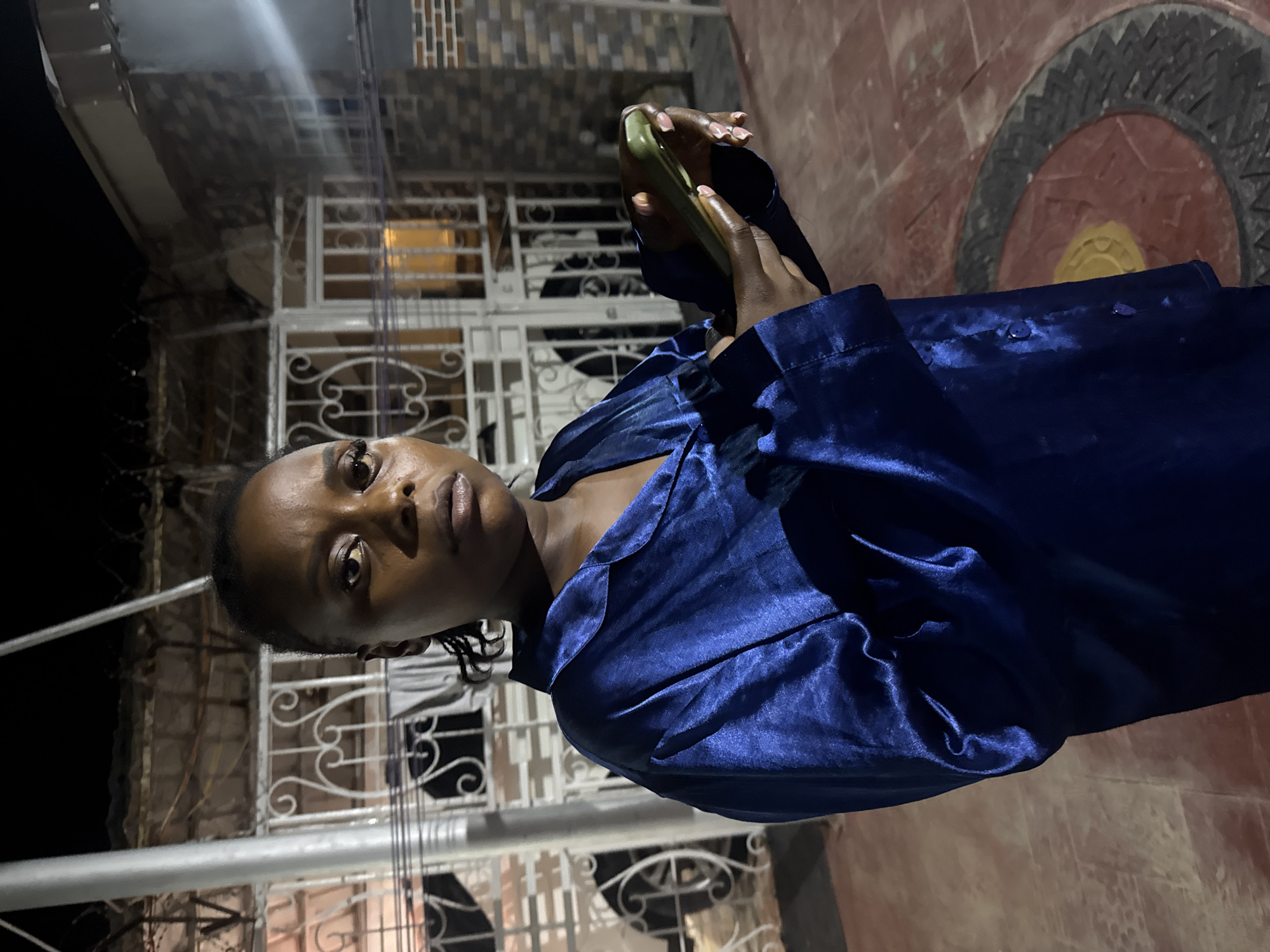A medium shot captures a young Black woman standing outdoors at night. She is wearing a shimmering, dark blue satin pajama top with a collar and buttons. The woman is holding a light green smartphone in her right hand, and her left hand is resting on the side of her body. Her hair is pulled back, and she has makeup on. Her expression is neutral. The background suggests a residential area in Abuja, Nigeria, with a metal gate, brick walls, and a tiled floor. A concrete pillar is visible on the left side of the frame, and barbed wire is strung along the top of the gate. The lighting indicates it is nighttime, with artificial light sources creating shadows.