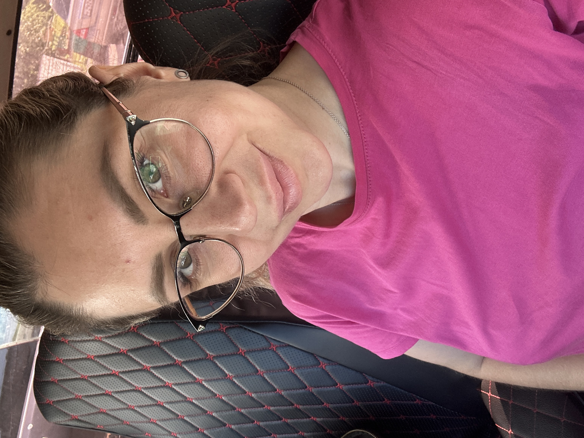 A woman with brown hair and green eyes, wearing a pink t-shirt and glasses, takes a selfie in the passenger seat of a car. The car's interior features black seats with red diamond stitching. The woman is also wearing a silver necklace and small silver earrings. In the background, a car is visible through the car window, suggesting an outdoor location in Novoaltaysk, Russia. The lighting suggests it is daytime.