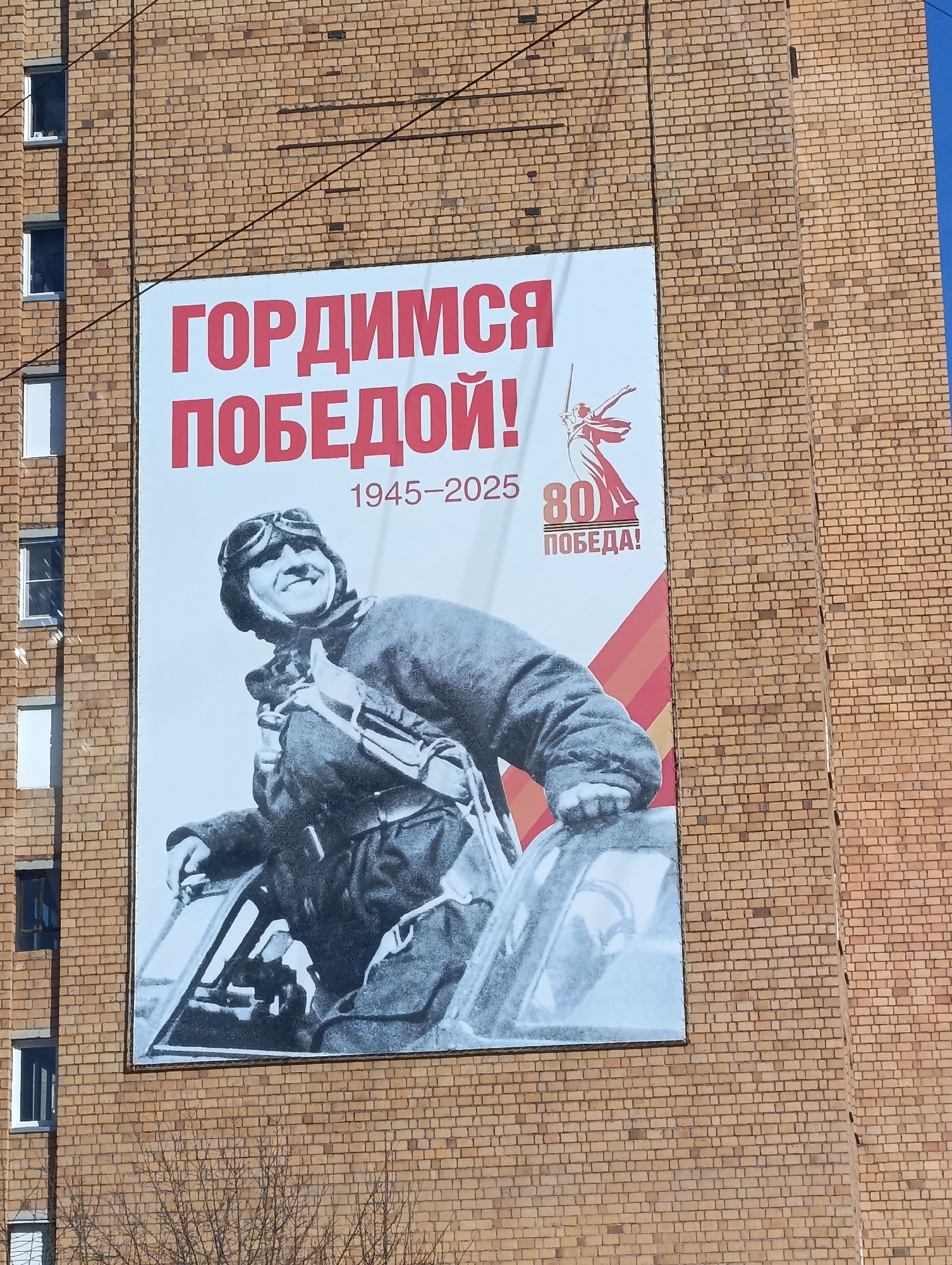 The image shows a large poster displayed on the side of a brick building. The poster commemorates the 80th anniversary of victory in World War II ("Pobeda!"). It features the phrase "Gordimsya Pobedyoy!" ("We are proud of the victory!") along with the years "1945-2025." An image of a Soviet aviator is featured prominently on the poster, along with a depiction of The Motherland Calls statue. Based on the design of the poster and the Cyrillic text, the image is likely located in Russia. Based on its distribution, this image is likely from the city of Krasnoyarsk, Russia.