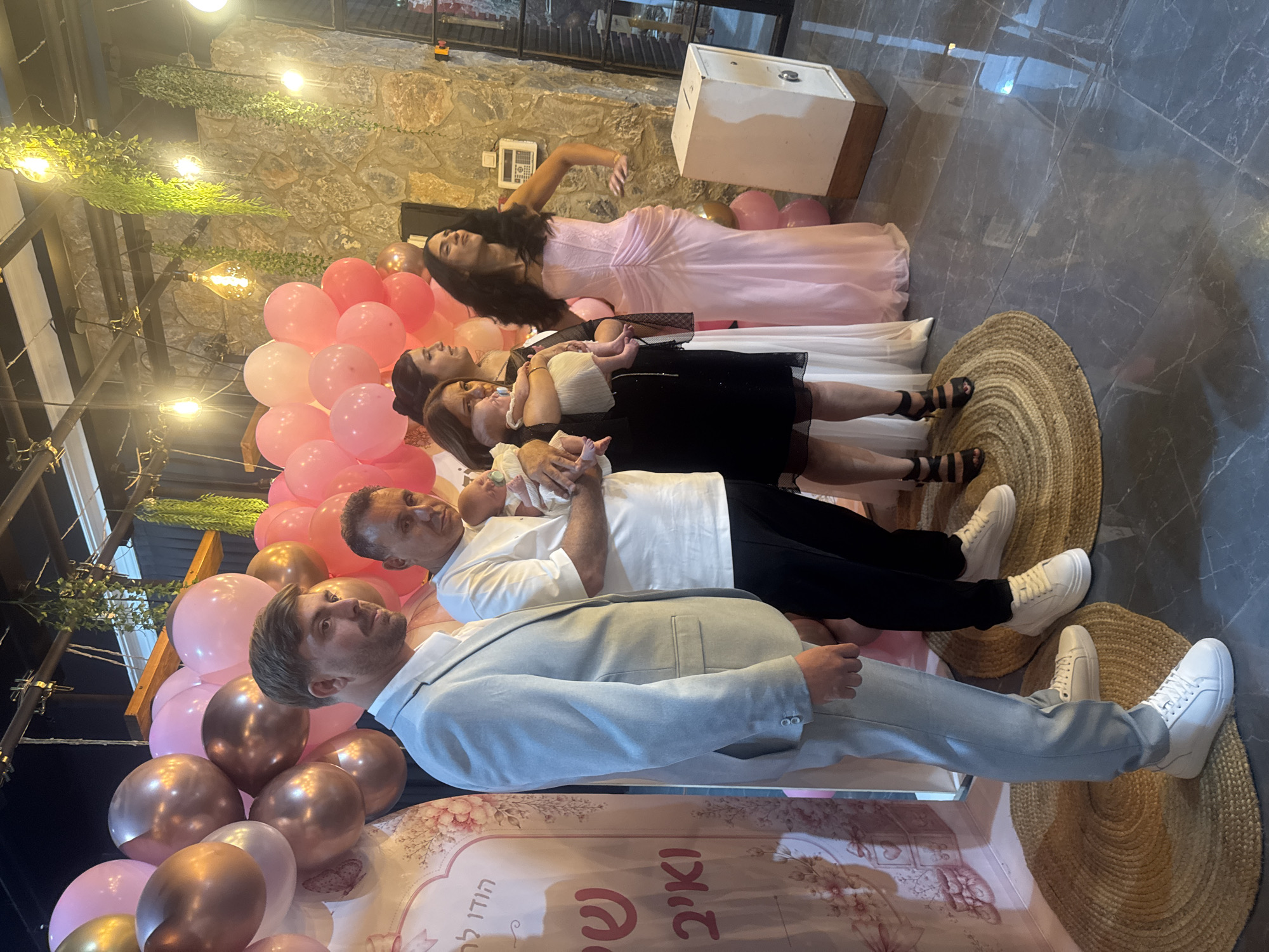 In Migdal HaEmek, Israel, a group of people is gathered for a celebration, likely a baby naming or similar event, as indicated by the pink and rose gold balloons and a decorative backdrop with floral motifs and Hebrew script.
On the left, a man in a light blue suit and white sneakers stands facing slightly away from the camera. To his right, another man in a white t-shirt and black pants holds a baby. In the center, a woman in a black dress holds another baby. Next to her, a woman in a white dress stands with her back to the camera. On the far right, a woman in a long, ruched pink dress with black heels poses with her hands on her hips.
The environment is indoors with a stone wall in the background and decorative lighting fixtures hanging from the ceiling, some with trailing greenery. A white safe-like object is positioned to the right of the group. The floor is tiled with a marbled pattern. The overall scene suggests a festive and intimate gathering.