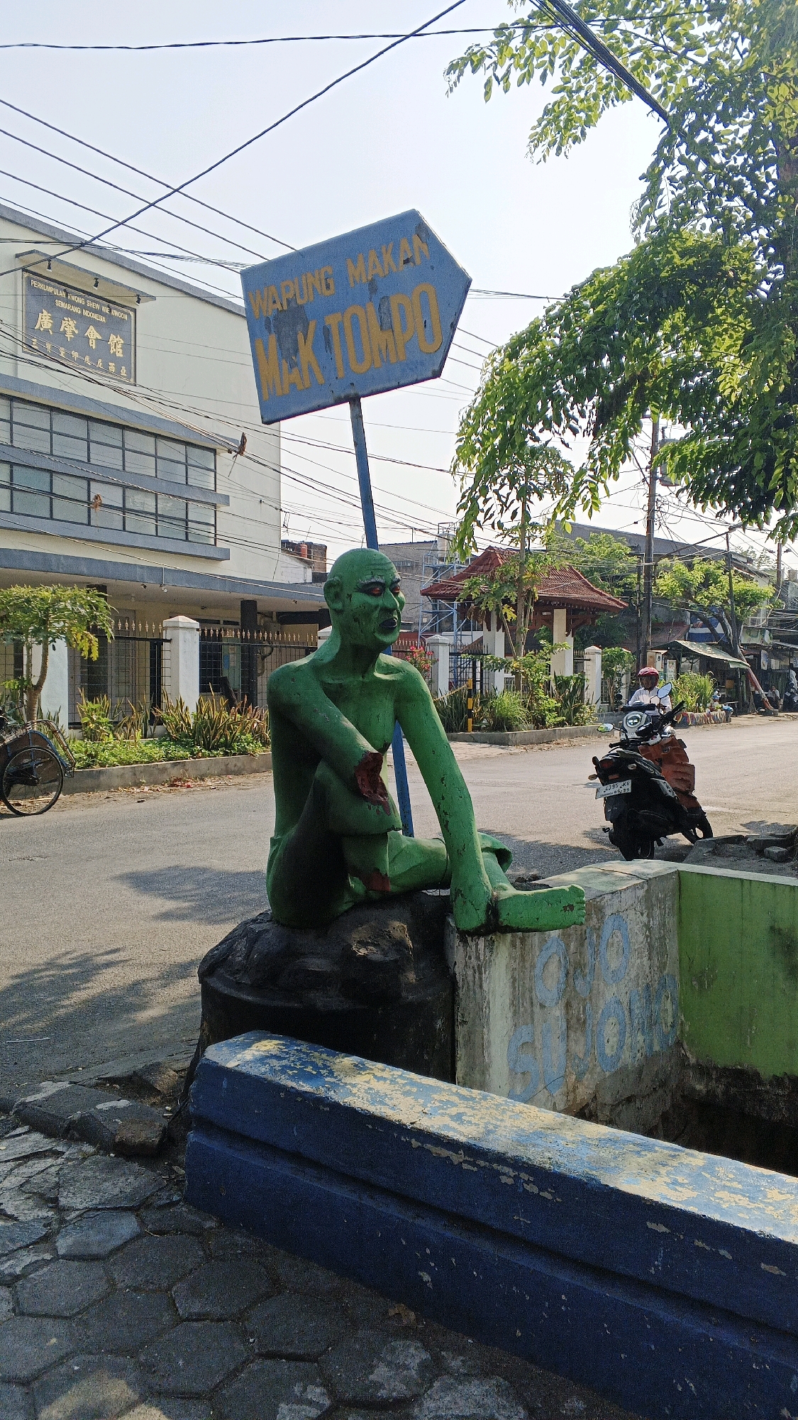 In Semarang, Indonesia, on a sunny day, a green statue of a seated figure with red eyes sits on a rock. The statue is positioned near a street, with a blue directional sign pointing to "WARUNG MAKAN MAK TOMPO". To the left of the statue, a building with a sign reading "PERKUMPULAN KHOO KHEW KHOON SEMARANG INDONESIA" is visible. A motorcycle with a rider passes by on the street. The foreground features a hexagonal patterned sidewalk and a blue structure. Lush green trees and vegetation are present throughout the scene, suggesting an outdoor urban environment.
