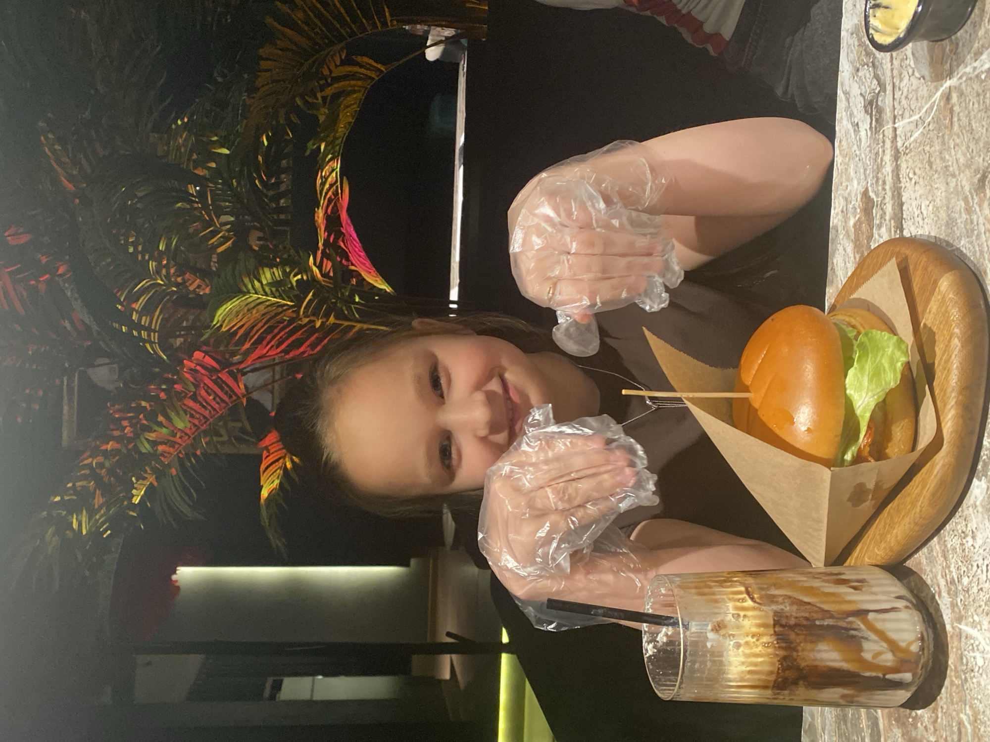 A young girl wearing plastic gloves sits at a table in a restaurant. A burger on a wooden plate and a partially consumed drink are on the table. A decorative plant is visible in the background. The location is the city of Chaikovsky, Russia.
