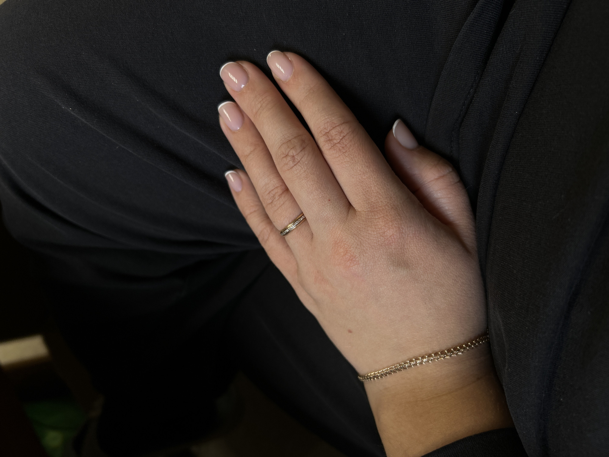 A photograph shows a woman's hand adorned with a gold ring on her ring finger and a delicate gold bracelet on her wrist. Her nails are manicured with a French tip design. The hand rests on a black fabric surface. There is no indication of location.
