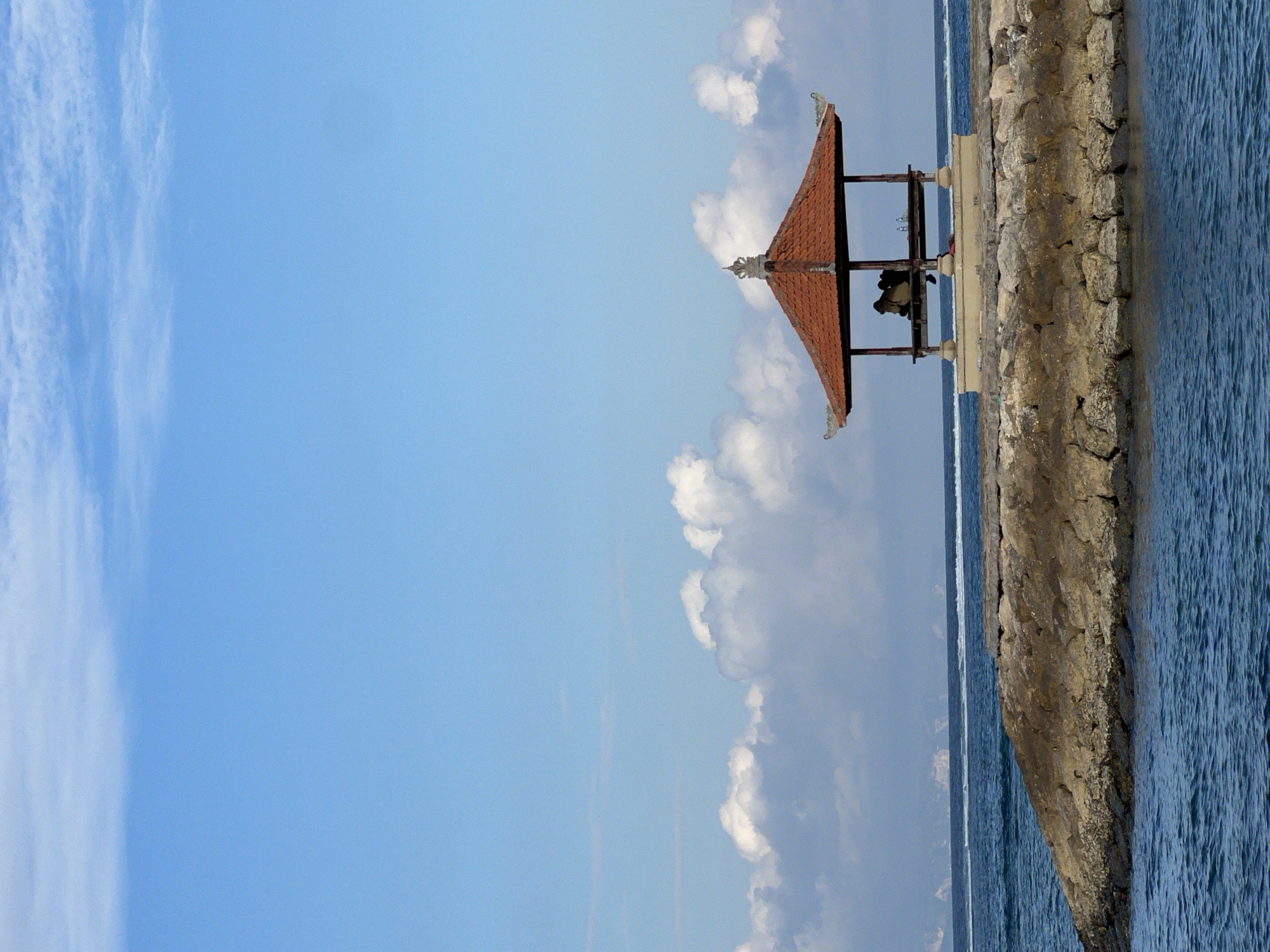 In Denpasar, Indonesia, a traditional Balinese pavilion with an orange tiled roof sits on a rocky embankment extending into the ocean. The pavilion is supported by pillars and houses two figures seated inside, facing away from the camera. Two bottles are visible on a shelf within the pavilion. The sky above is a clear blue, with scattered white clouds forming near the horizon. The ocean is a deep blue, with gentle waves lapping against the rocks. The scene evokes a sense of tranquility and a typical coastal view in Denpasar.