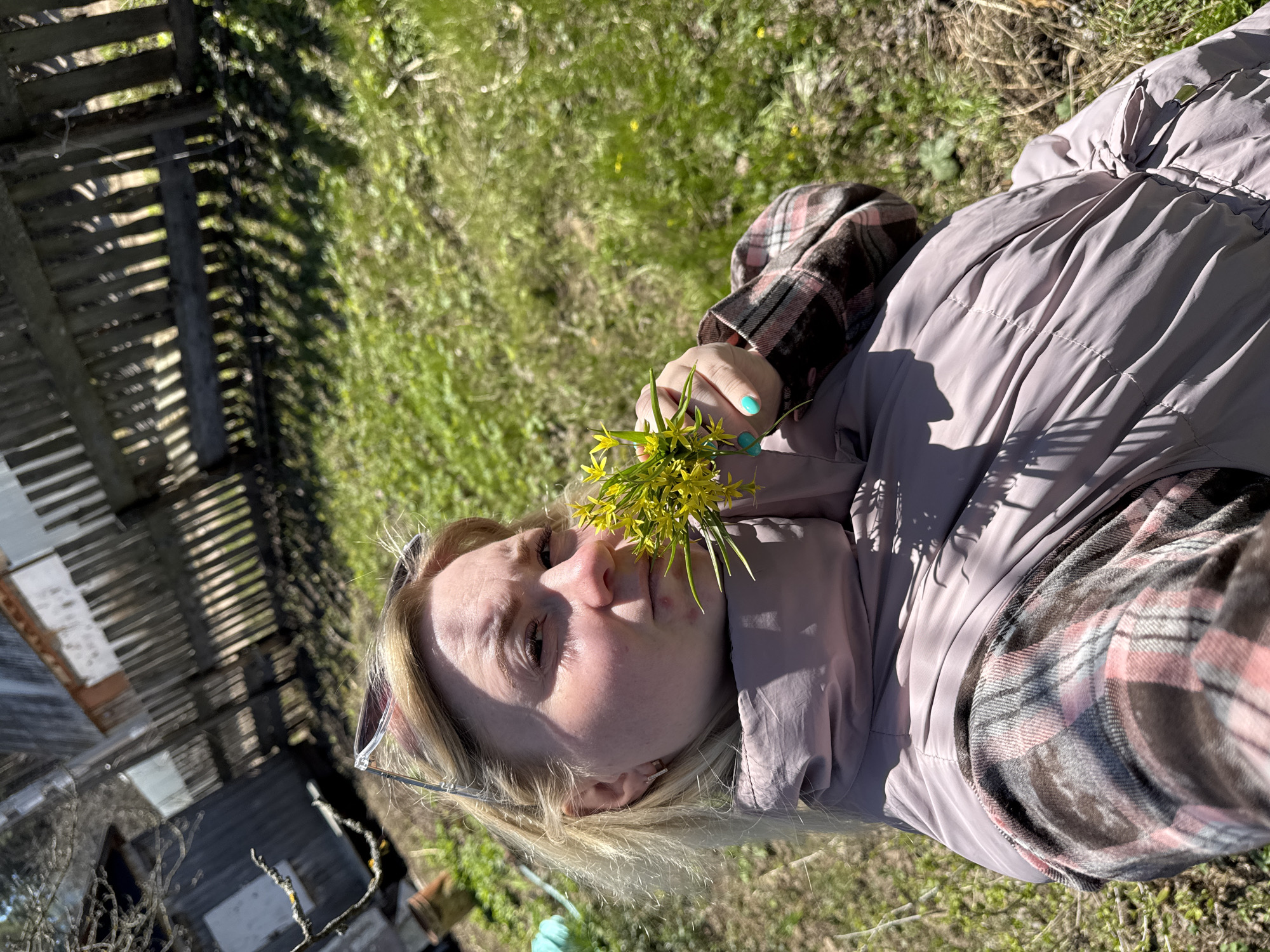 A light-skinned person with blonde hair, wearing a pale purple quilted jacket over a pink and grey plaid shirt, is lying on grass. They are holding a cluster of small yellow flowers near their face. Sunglasses are perched on their head. To the left, there's a blurred view of a wooden fence and a building. The setting appears to be outdoors, possibly in Chaikovsky, Russia.