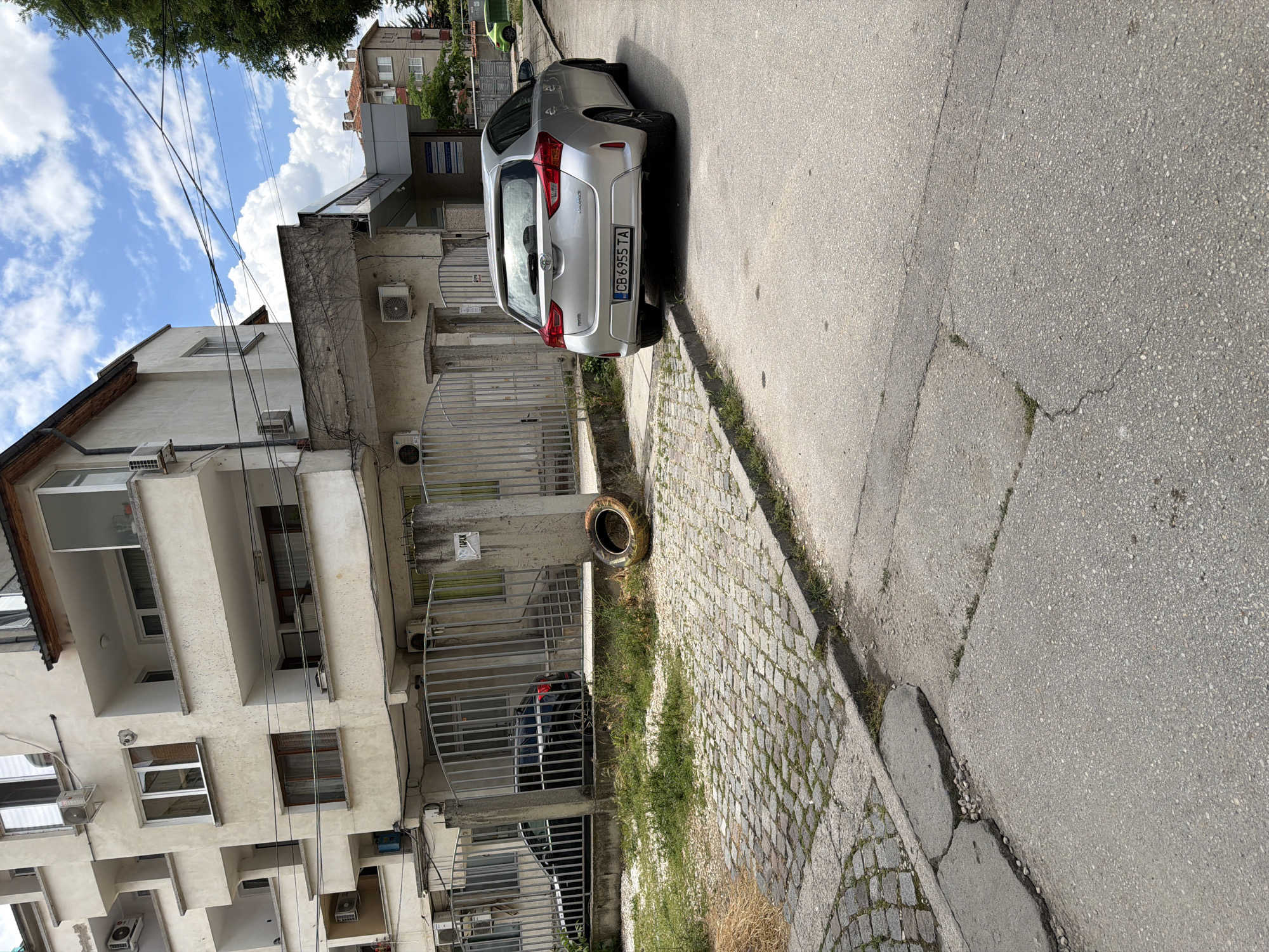 This is a street-level, eye-level, medium shot captured on a cloudy day in Pazardzhik, Bulgaria. The scene features a silver Toyota hatchback parked on the right side of a paved road. The car's license plate reads "CB 6955 TA". To the left of the car and the road is a cobblestone pathway bordered by a concrete curb, which appears cracked in places. Beyond the cobblestones is a patch of overgrown grass and a discarded tire. Behind the grass and tire, a grey metal fence encloses a paved area. Through the fence, a dark-colored SUV is partially visible.
The background is dominated by a multi-story apartment building with cream-colored walls and numerous balconies. Some balconies have visible air conditioning units. Further in the background, another residential building with red-tiled roofs and white window frames can be seen, along with trees. Overhead, power lines crisscross against a sky filled with white cumulus clouds. A small sign with Cyrillic text, possibly indicating "GARAGE," is affixed to a concrete post near the fence. The overall impression is one of a moderately urban residential area.