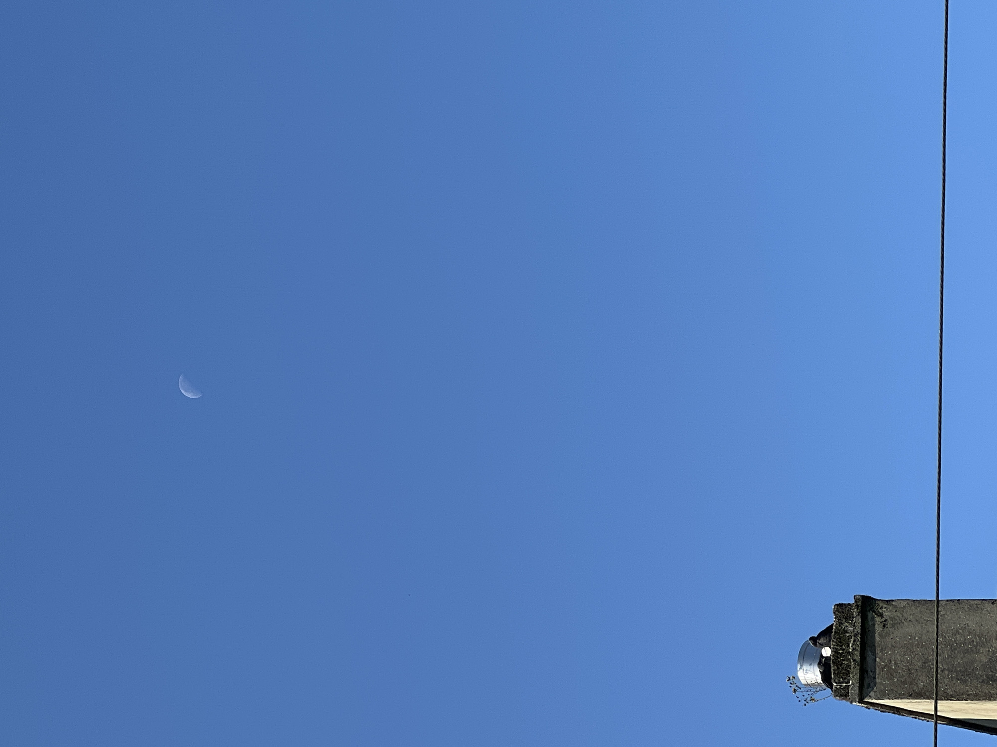 The image shows a clear blue sky above a chimney in Mountcharles, Ireland. A crescent moon is visible in the upper portion of the sky. On top of the chimney, there is a silver cylindrical object and two birds are perched on it. A thin wire runs horizontally across the lower part of the image.