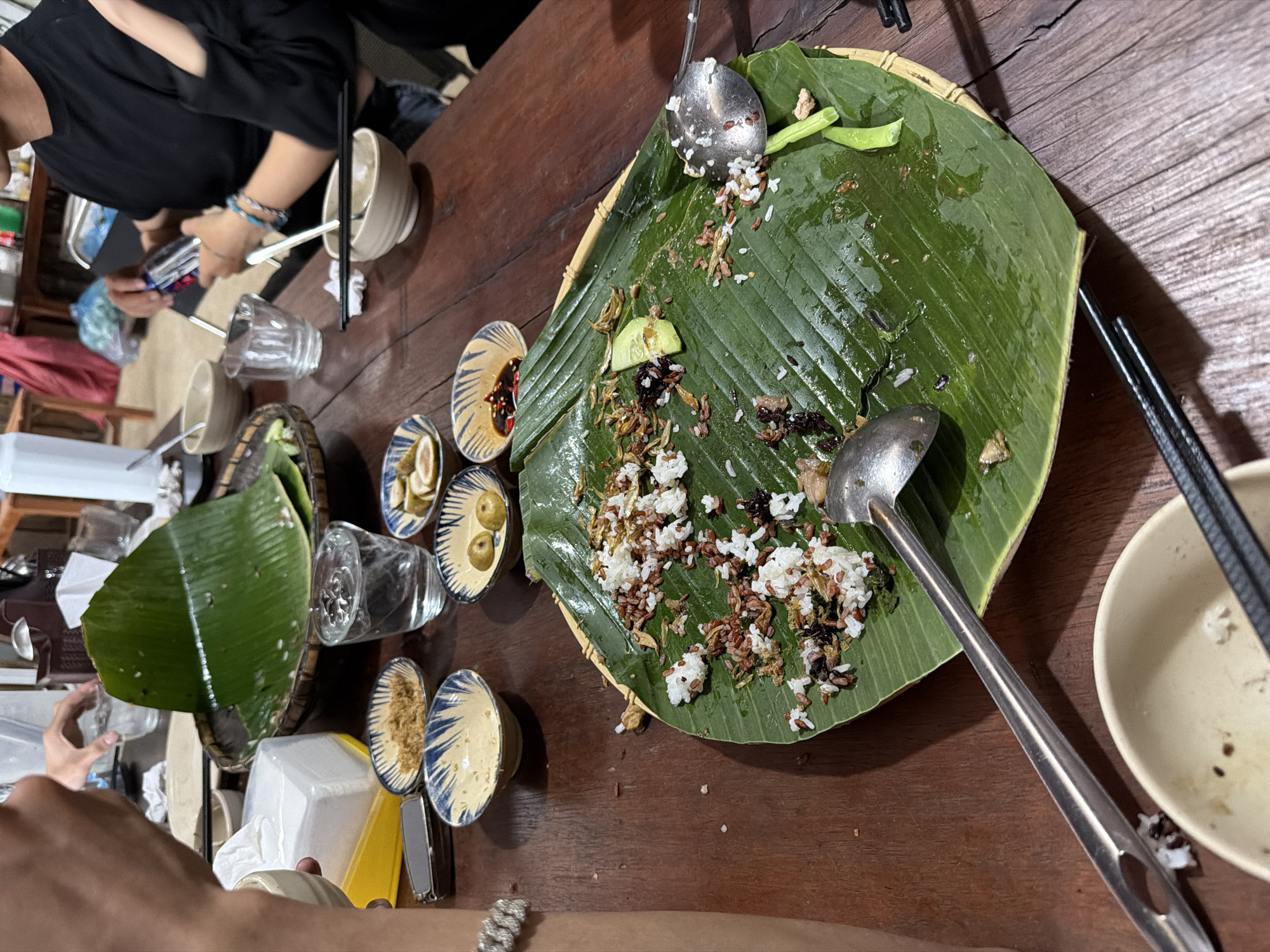The image depicts an indoor, communal dining scene, likely in Măng Đen, Vietnam. The setting is a dark wooden table cluttered with the remnants of a meal.
At the center, a large, vibrant green banana leaf serves as a platter, placed on a woven rattan or bamboo tray. It holds a mixture of partially eaten food, including white and brown rice, small fried fish or insects, sliced cucumber, and other green vegetable pieces, along with some dark, unidentified ingredients. Metal serving spoons rest on the leaf.
Surrounding the main dish are several small, blue-and-white patterned ceramic bowls containing various condiments and side dishes. These include dark sauces (possibly chili or fish sauce), sliced lemons or limes, and granular toppings that might be fried shallots or other spices. Empty and partially filled ceramic bowls, glass cups, black chopsticks, and lighter-colored individual spoons are also scattered across the table. Other objects include a white napkin dispenser and a yellow plastic container.
A person wearing a black top and blue bracelets is partially visible on the left side of the frame, holding what appears to be a smartphone or a small item. Another person's arm, adorned with a silver bracelet, is visible in the bottom right corner. The scene suggests a casual gathering, with the meal nearing its end. The lighting indicates an indoor setting, possibly an evening meal. The traditional serving style on a banana leaf and the types of food are consistent with local cuisine in Vietnam. No visible text is clearly legible in the image.
