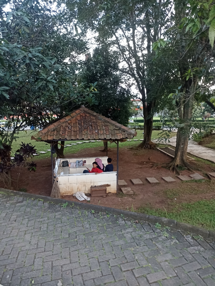 The image shows a small, tiled structure with a tiled roof, located in a park-like setting in Jatinangor, Indonesia, at the coordinates -6.931 latitude and 107.776 longitude. The structure is situated on a grassy area, surrounded by trees and bushes. The image is taken from a slightly elevated vantage point, looking down at the structure. Two individuals are sitting inside the structure. One is a young woman wearing a red shirt and a hijab, sitting on a bench with her back to the camera. Next to her, a person in a blue shirt is sitting, partially obscured. There is a small bench between them. A few stepping stones are visible in front of the structure, leading toward a paved pathway. The environment is green and lush. The sky is overcast.