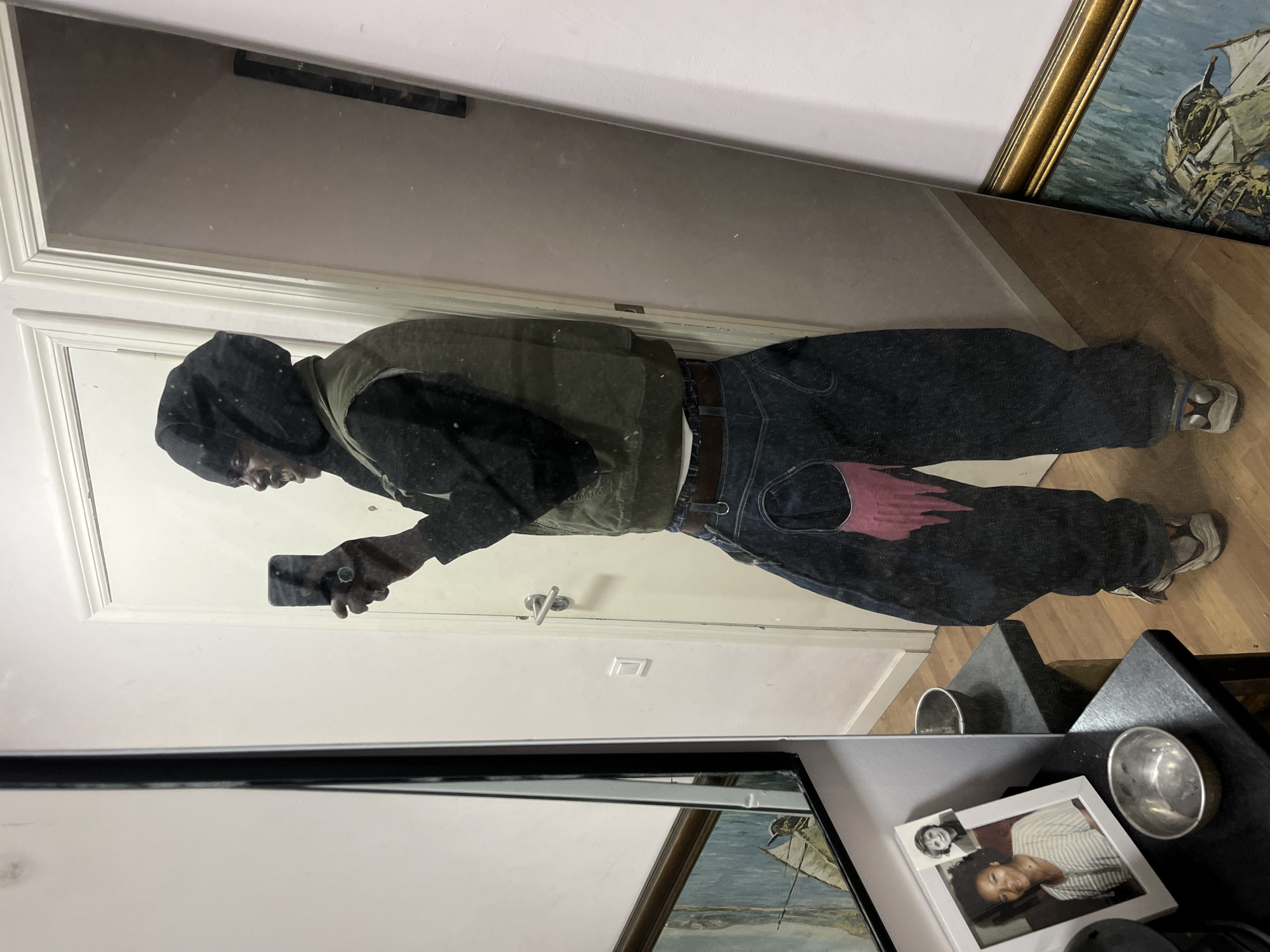 A person wearing a dark hooded sweatshirt, a green vest, and dark pants with a red design is reflected in a mirror while holding a cell phone. The person appears to be taking a selfie. The mirror shows the reflection of a door, a painting, a wooden floor, and a table with a picture frame and a small metal bowl on it. The setting is a room with light walls and a door. The location in the city of Barcelona, Spain cannot be determined from the provided image.