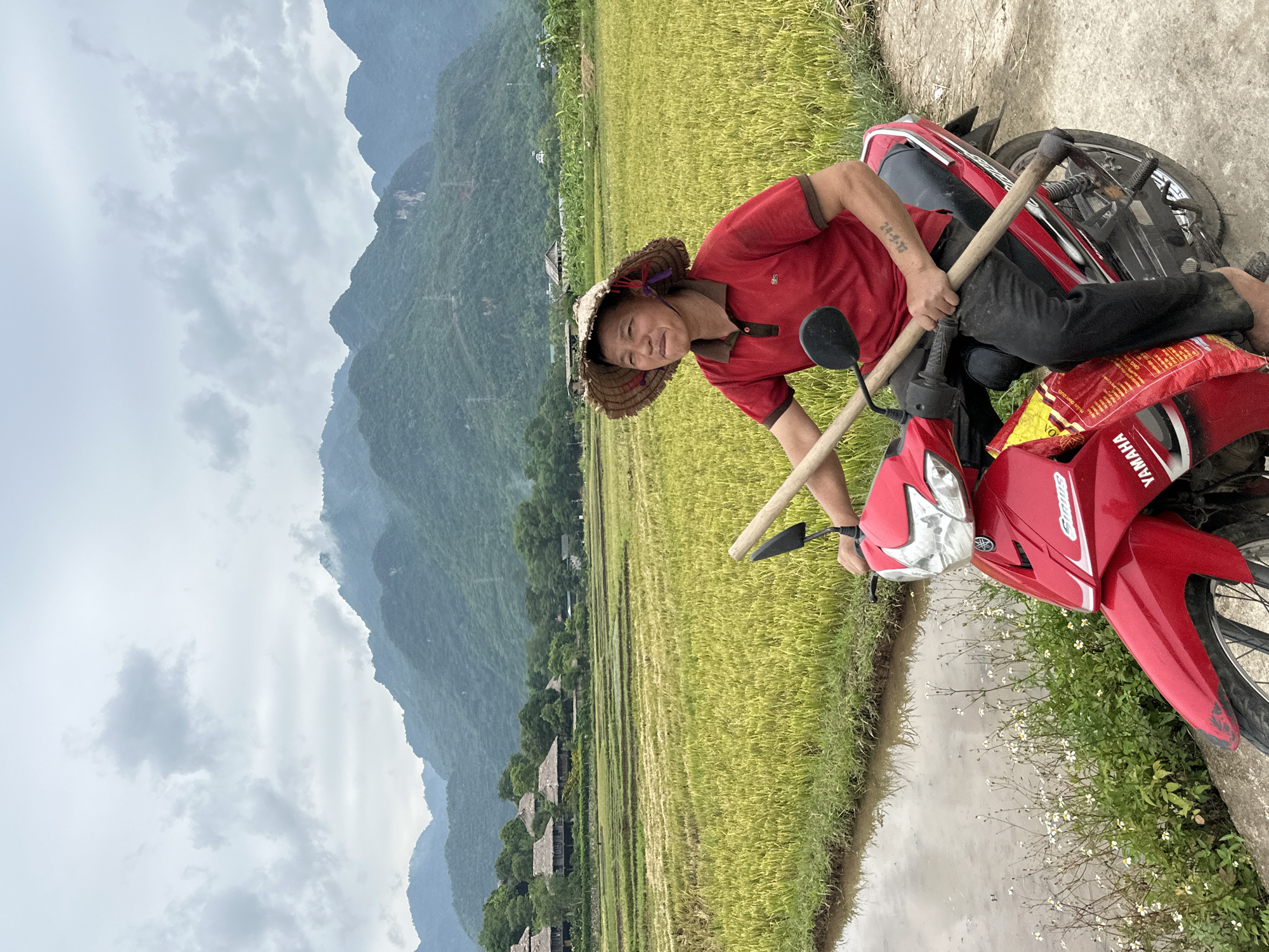 A person wearing a conical straw hat and a red shirt sits astride a red Yamaha motorcycle on a dirt path next to a flooded rice paddy. The person is holding a wooden pole with a dark-colored tool attached to one end. The background features lush green rice fields stretching towards a village of stilted houses with thatched roofs. Beyond the village, rolling green mountains are shrouded in a light mist under a cloudy sky. The person's left arm bears a tattoo. A yellow and red bag is strapped to the rear of the motorcycle. The image appears to have been taken in Mai Chau, Vietnam, given the characteristic landscape and architecture.
